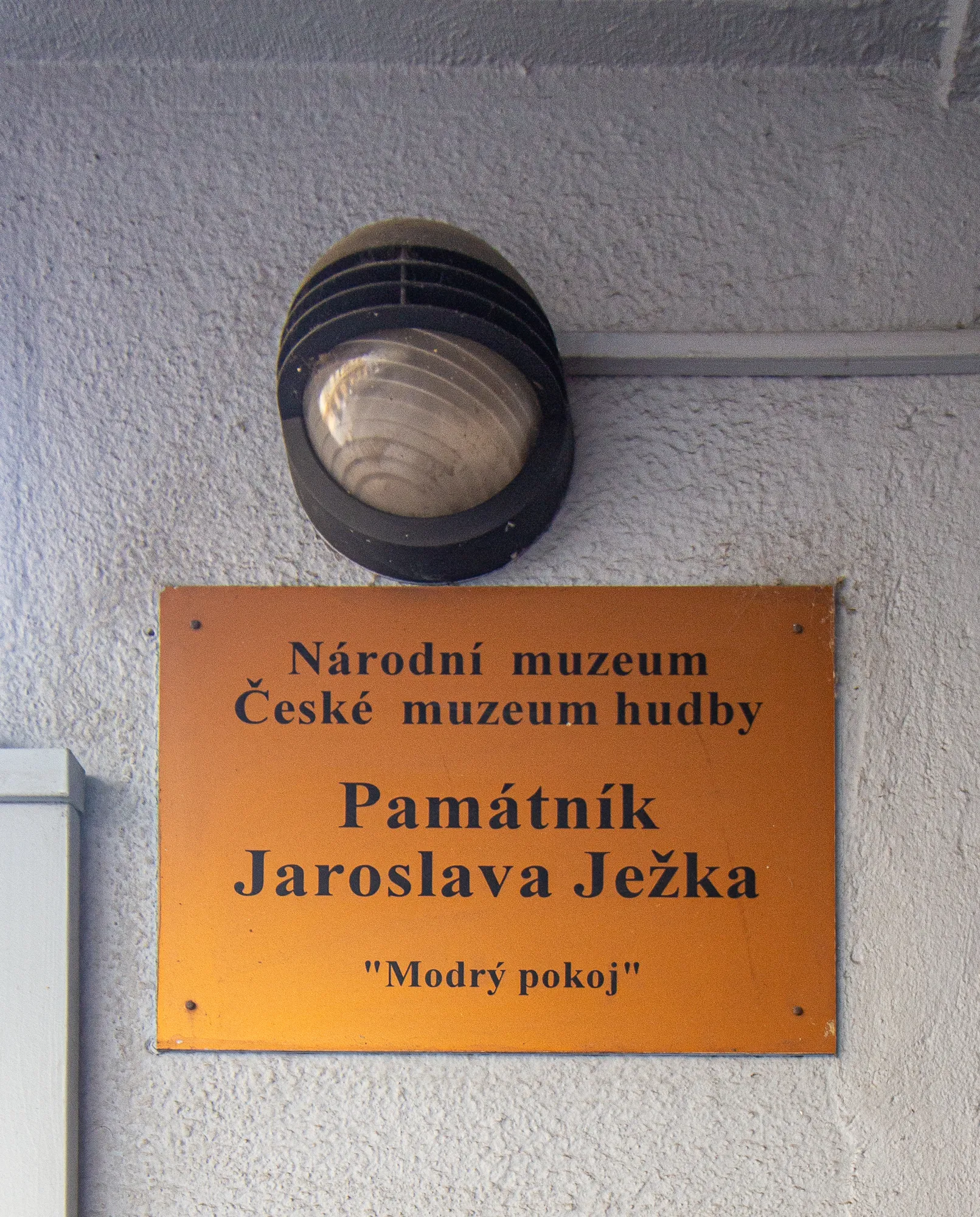 Pomarańczowy znak na teksturowanej ścianie z napisem 'Národní muzeum České muzeum hudby Památník Jaroslava Ježka 'Modrý pokoj''