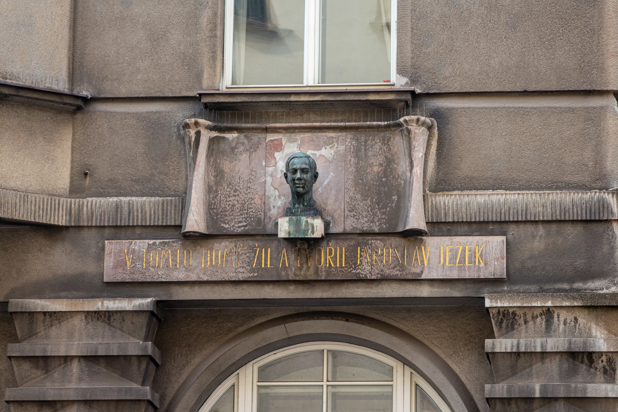 Brązowe popiersie Jaroslava Ježka na płycie pamiątkowej zamontowanej na fasadzie budynku z wyrytymi poniżej napisami.