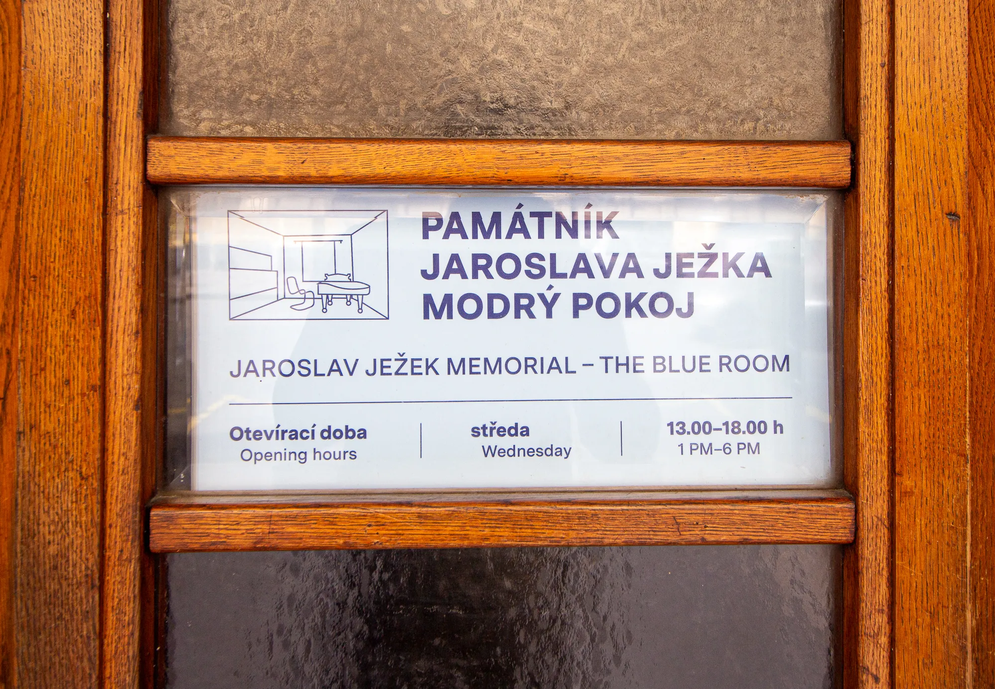 Drewniane drzwi ze szklanym panelem z napisem 'PAMÁTNÍK JAROSLAVA JEŽKA MODRÝ POKOJ' i 'JAROSLAV JEŽEK MEMORIAL - THE BLUE ROOM' oraz godzinami otwarcia.