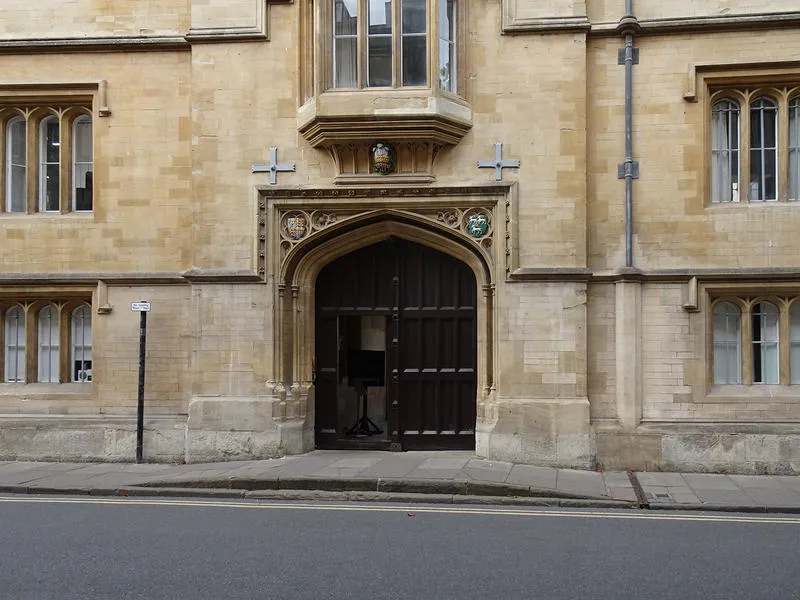 Vista exterior del Jesus College que muestra una puerta de entrada arqueada de madera en una fachada de piedra color miel con ventanas góticas y un escudo de armas sobre la puerta.