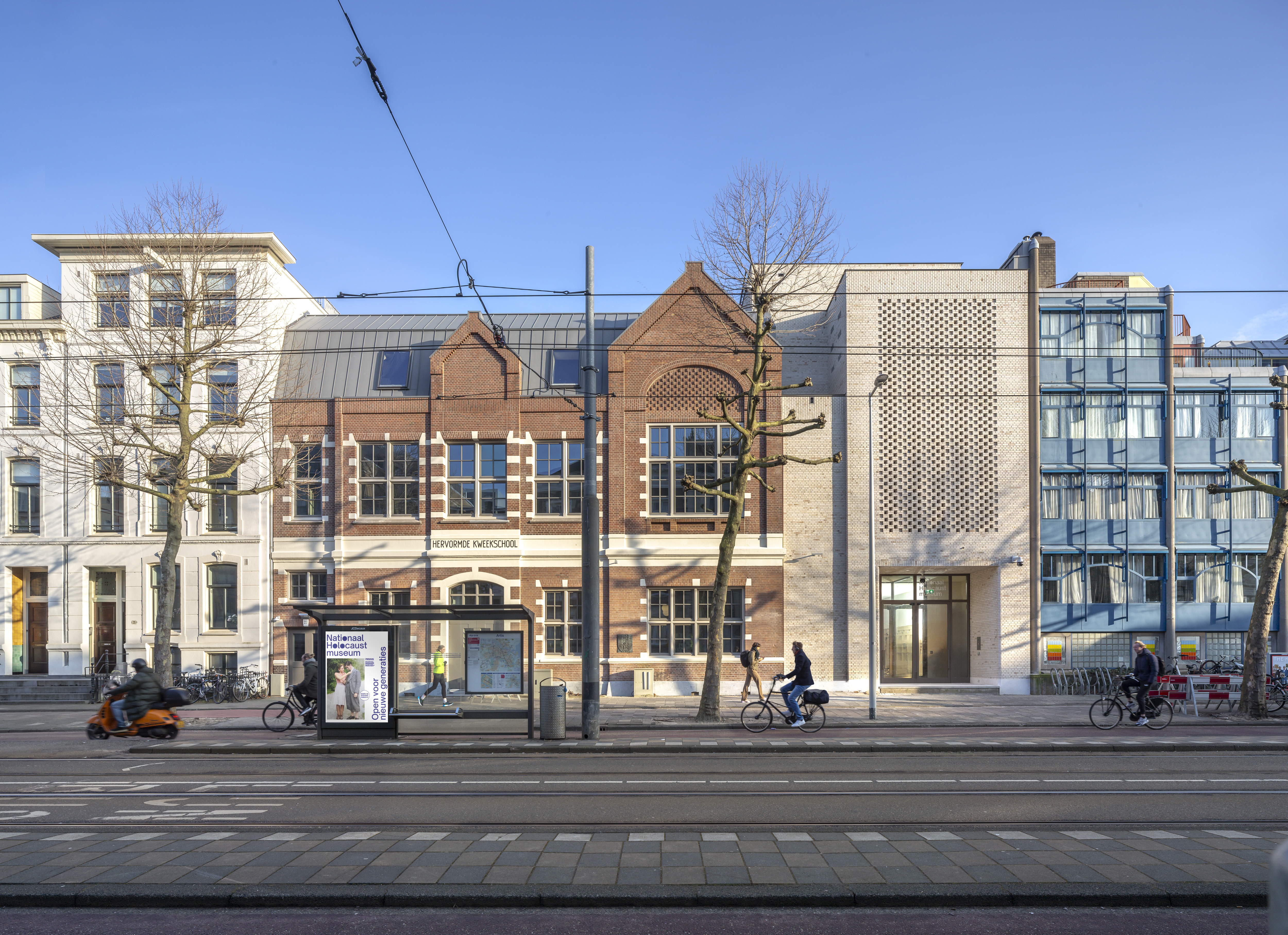 Hollandsche Schouwburg building with red brick facade on a city street, tram tracks, bicycles, and pedestrians under clear blue sky