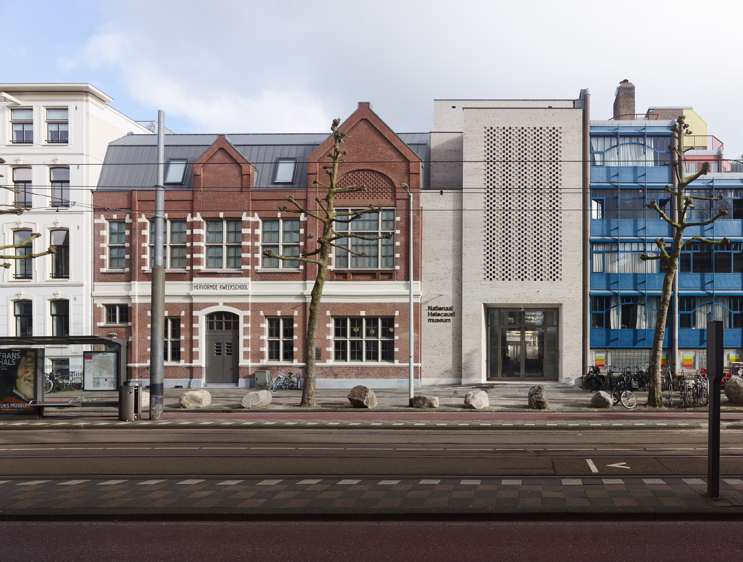 Street view of historic red-brick building with gabled roof alongside modern concrete structure with patterned facade, tram tracks in foreground.