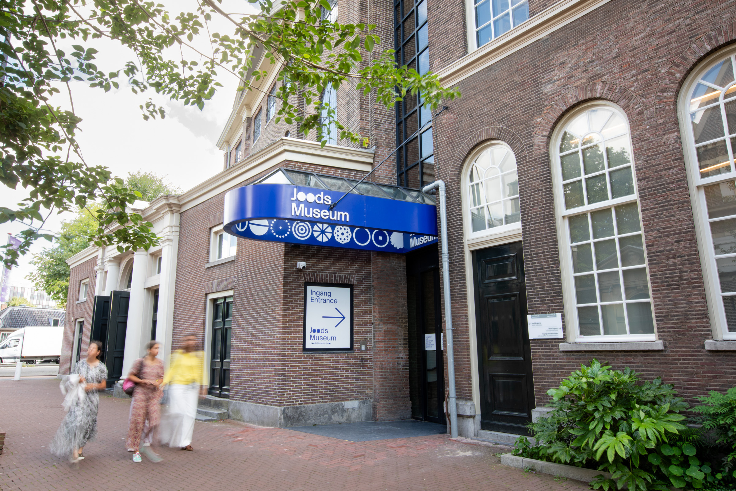 Wejście do Muzeum Żydowskiego z niebieskim zadaszeniem, ceglany budynek z łukowymi oknami, ludzie spacerujący po chodniku w Amsterdamie
