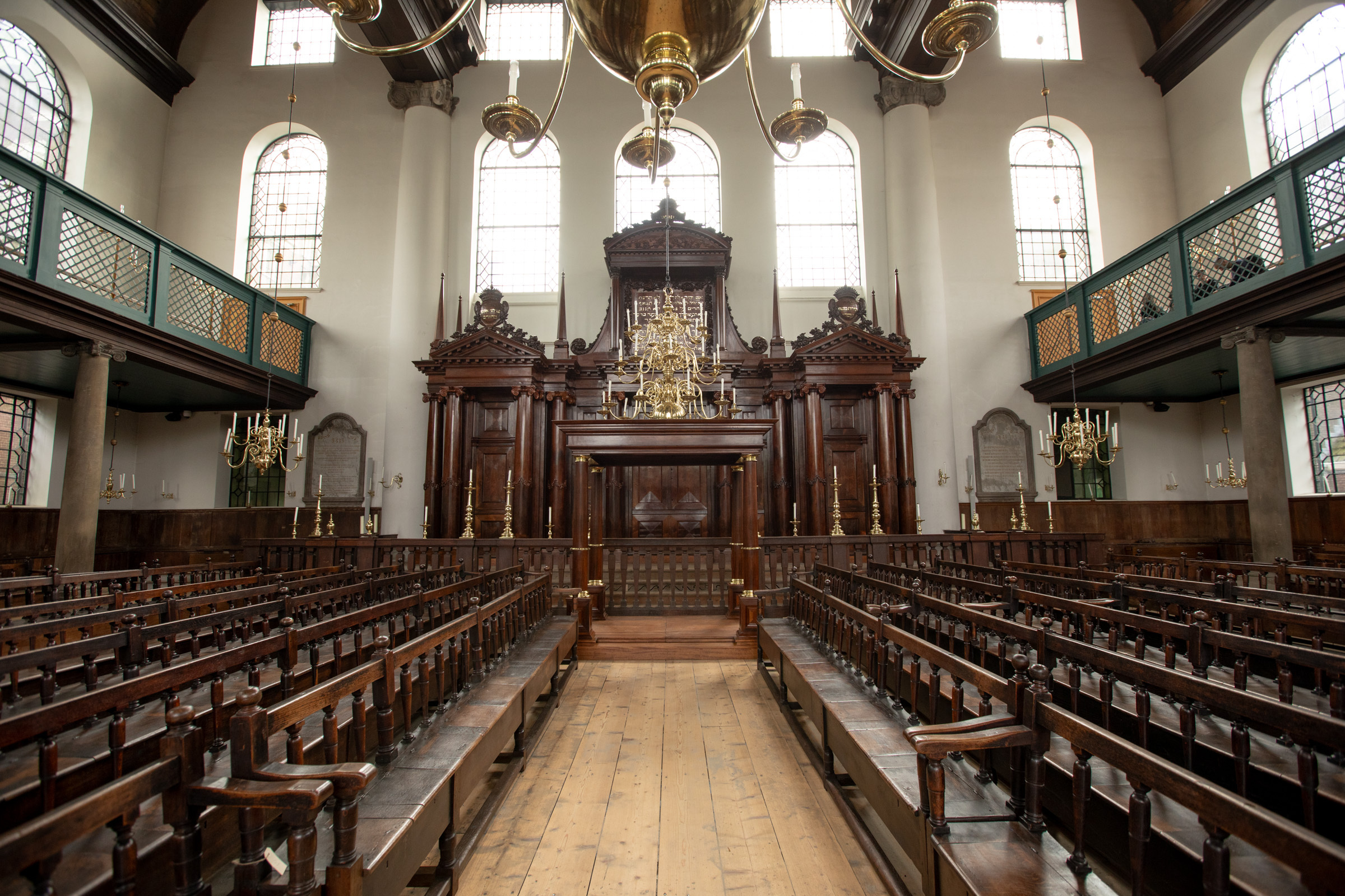 Drewniane ławki ustawione są w stronę ozdobnej arki pod wysokimi sufitami z dużymi oknami we wnętrzu zabytkowej synagogi.