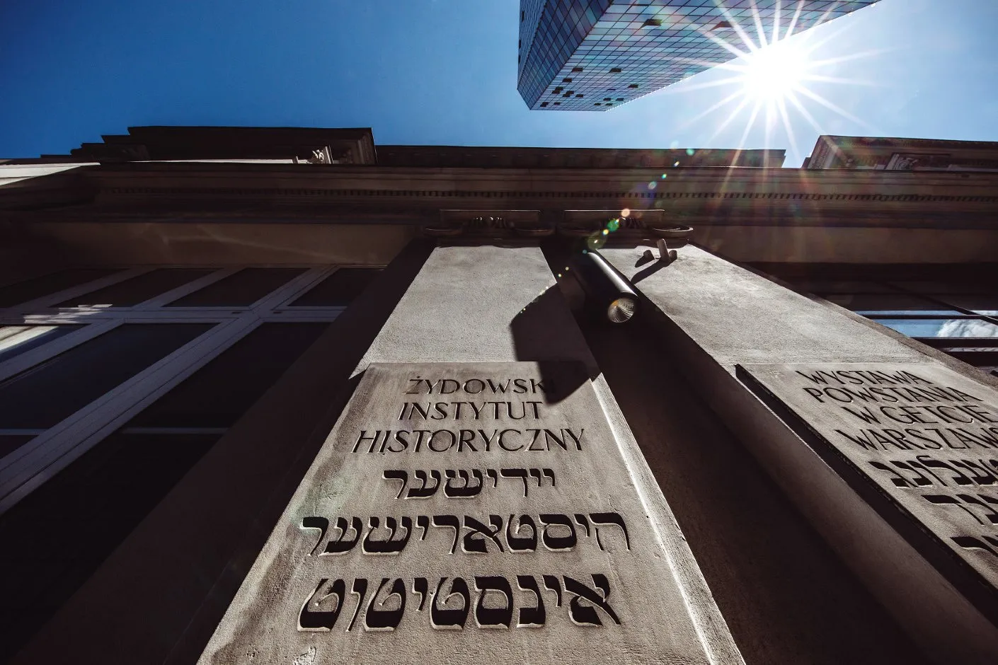Stone facade of the Jewish Historical Institute with inscriptions in Polish, Hebrew, and Yiddish, under a bright blue sky with sun flare.