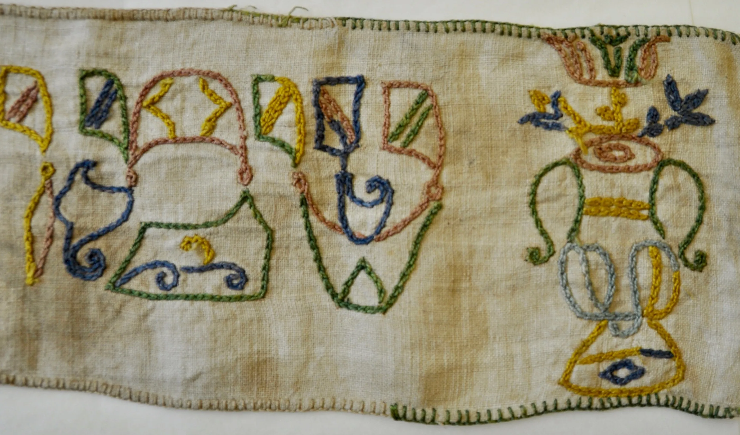 Geborduurd linnen textiel met kleurrijke Hebreeuwse letters en decoratieve bloemmotieven in blauw, groen, geel en rood garen op een beige stoffen achtergrond.