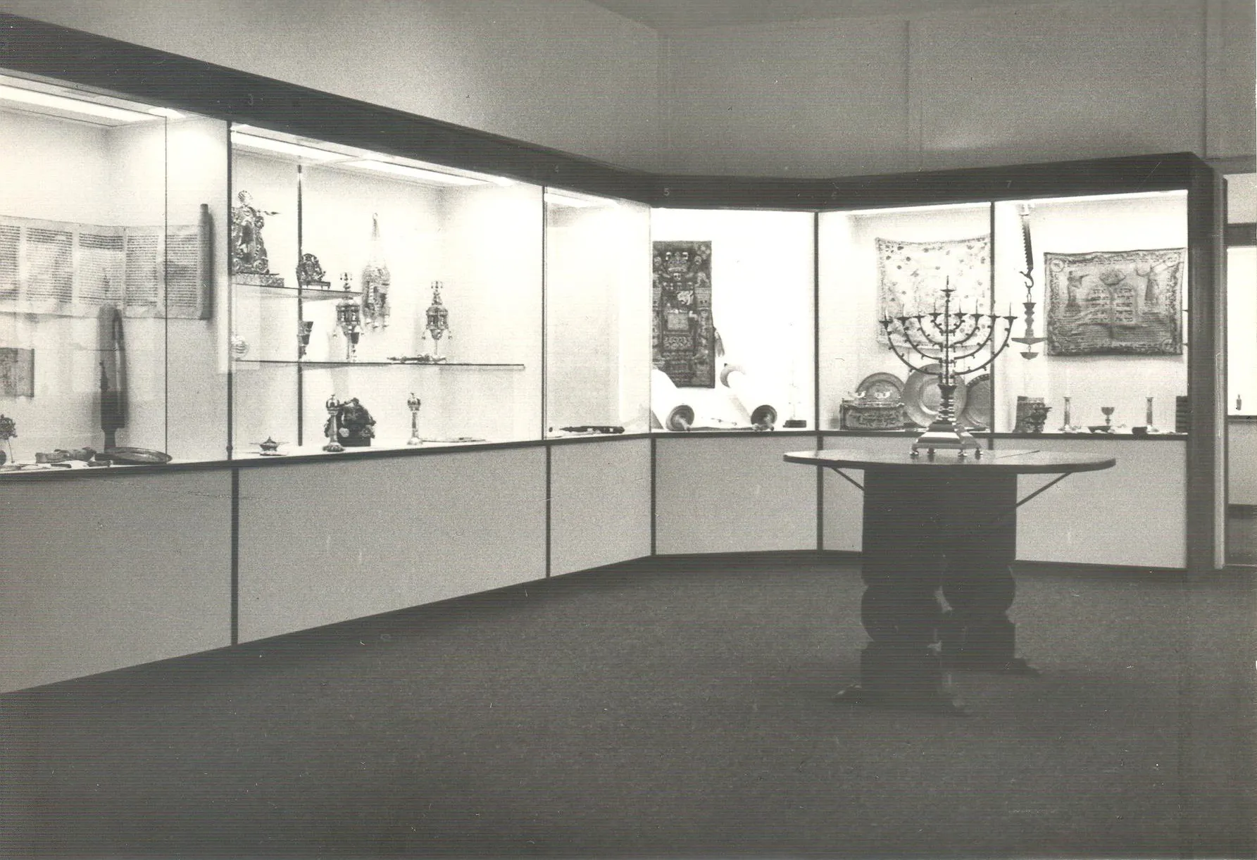 Fotografía en blanco y negro de una sala de exposiciones de un museo con vitrinas a lo largo de paredes curvas y una mesa central que muestra una menorá y otros artefactos ceremoniales judíos.
