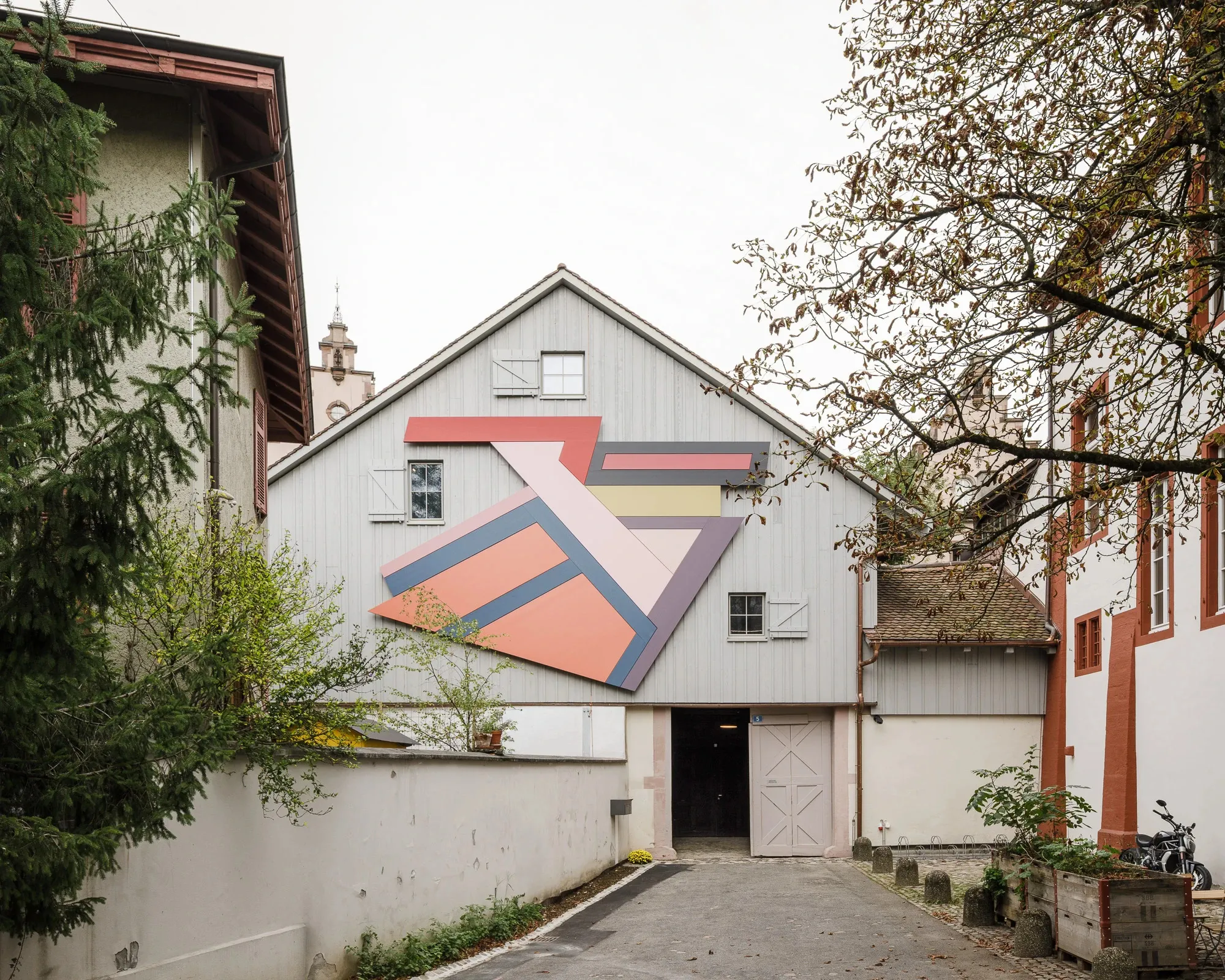 Wit historisch graanpakhuis met een grote kleurrijke geometrische muurschildering op de gevel, gezien vanuit een smal kasseiensteegje met bomen aan weerszijden