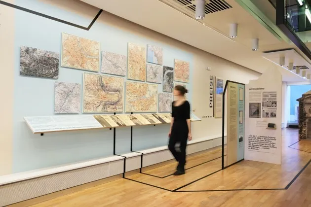 Een vrouw loopt door een tentoonstellingsruimte van een museum met ingelijste historische kaarten en documenten aan een witte muur, vergezeld door displaystandaarden en een informatiepaneel op een lichte houten vloer.