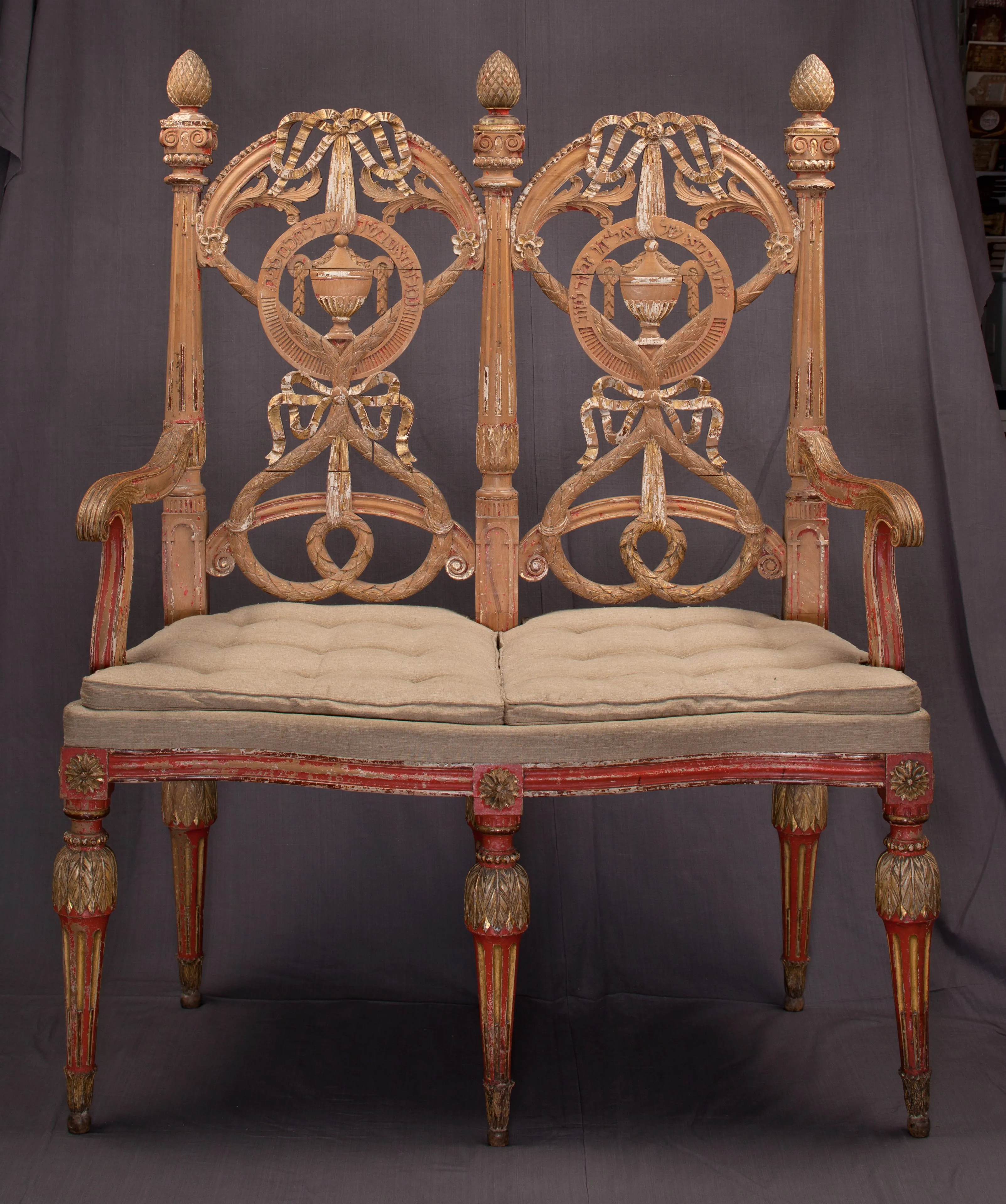 Rijkversierde houten tweezitsbank met uitgesneden rugleuningen met urnmotieven en rood met goud beschilderde poten, geïdentificeerd als een Brit milah-bank.