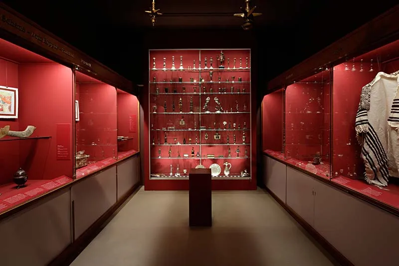 Museumgalerie met glazen vitrines met rode achterwanden langs beide muren, gevuld met ceremoniële voorwerpen, en een hoge verlichte vitrine aan het uiteinde met een voetstuk in het middenpad.