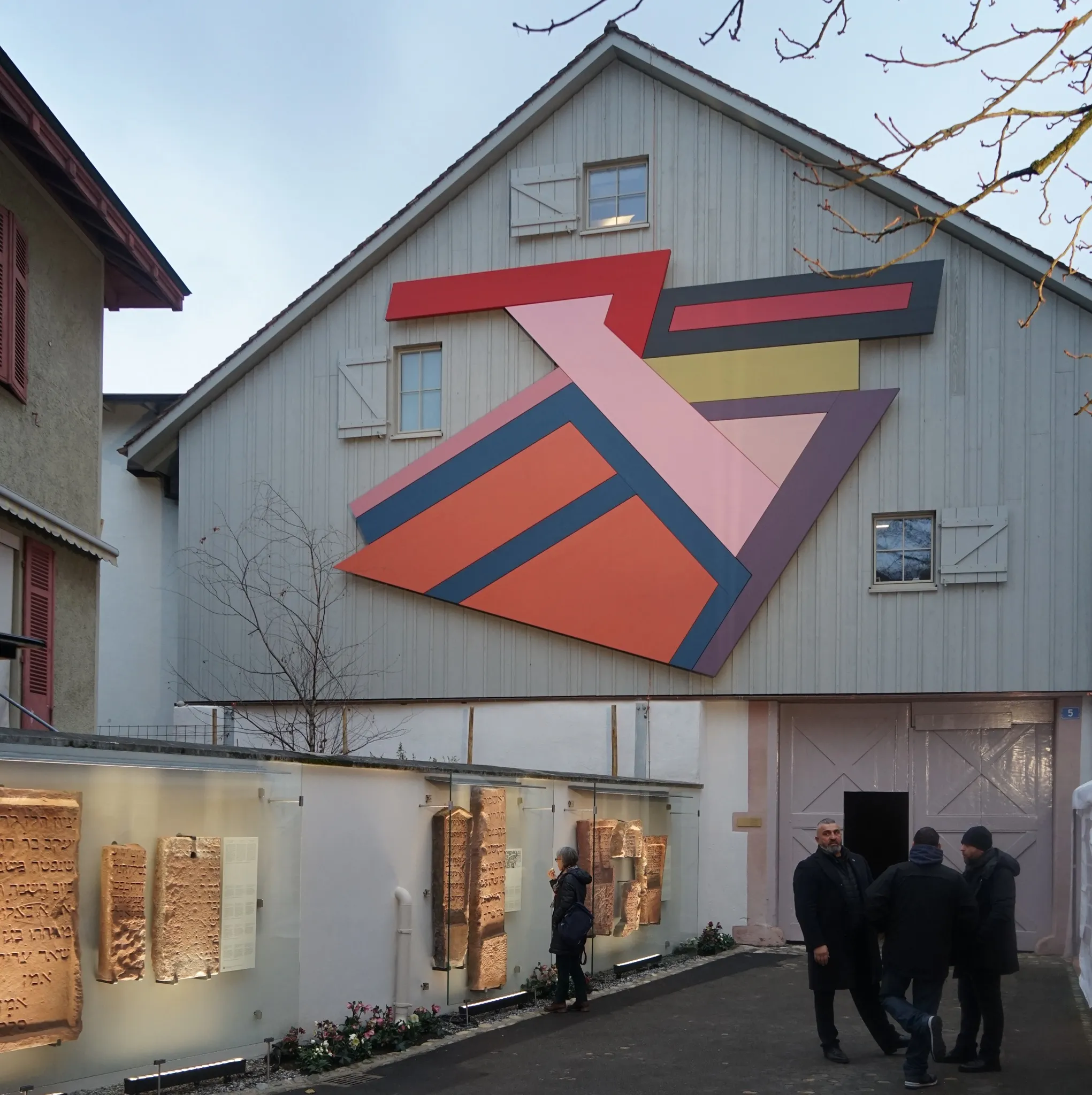 Buitenkant van het Joods Museum van Zwitserland met een grote kleurrijke geometrische muurschildering op de grijze gevel van het gebouw, met verlichte displaypanelen langs een binnenplaatsmuur en bezoekers die in de buurt staan.