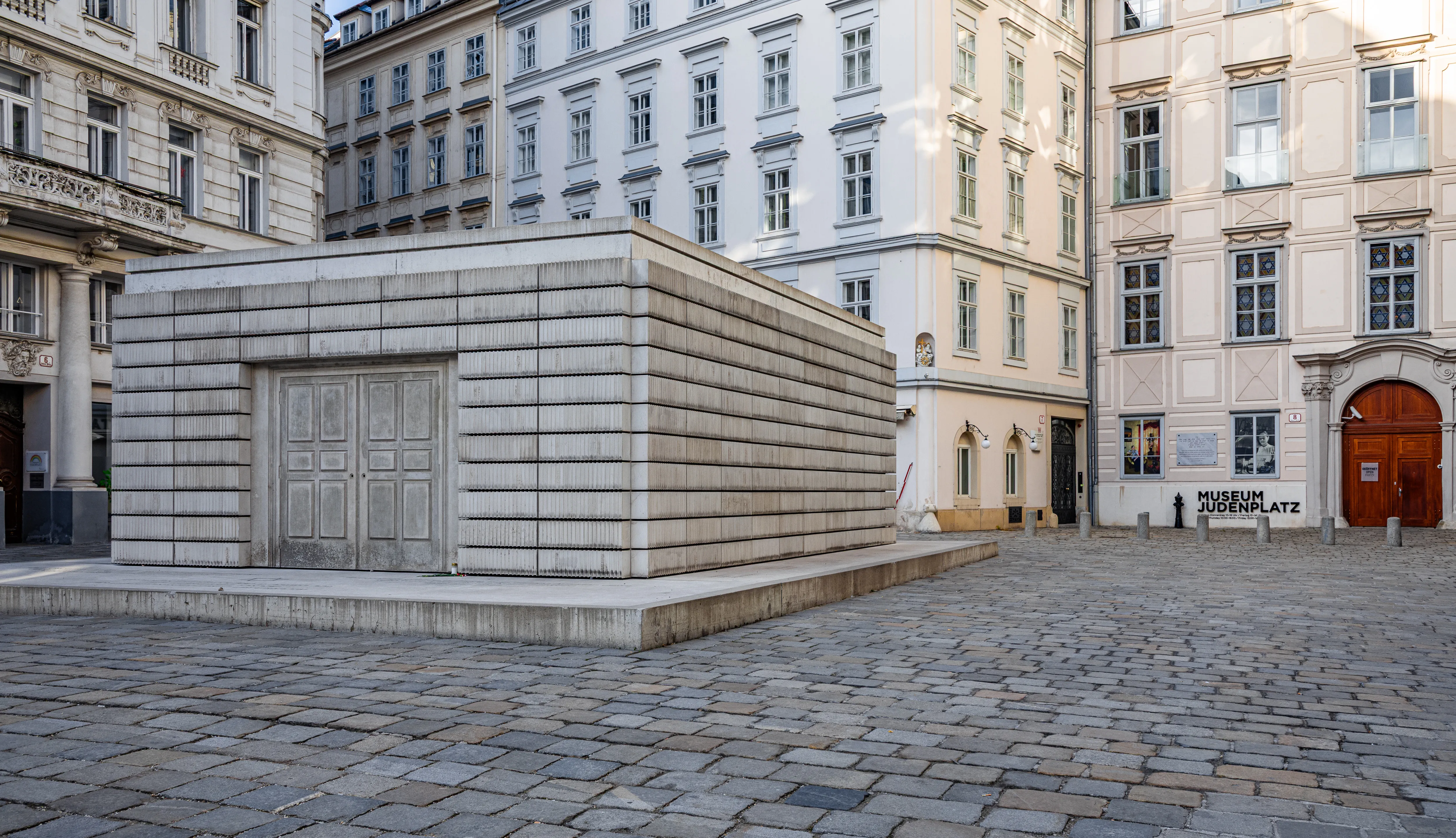 Een modern stenen kubusvormig monument op een geplaveide plein, omringd door klassieke Europese gebouwen.