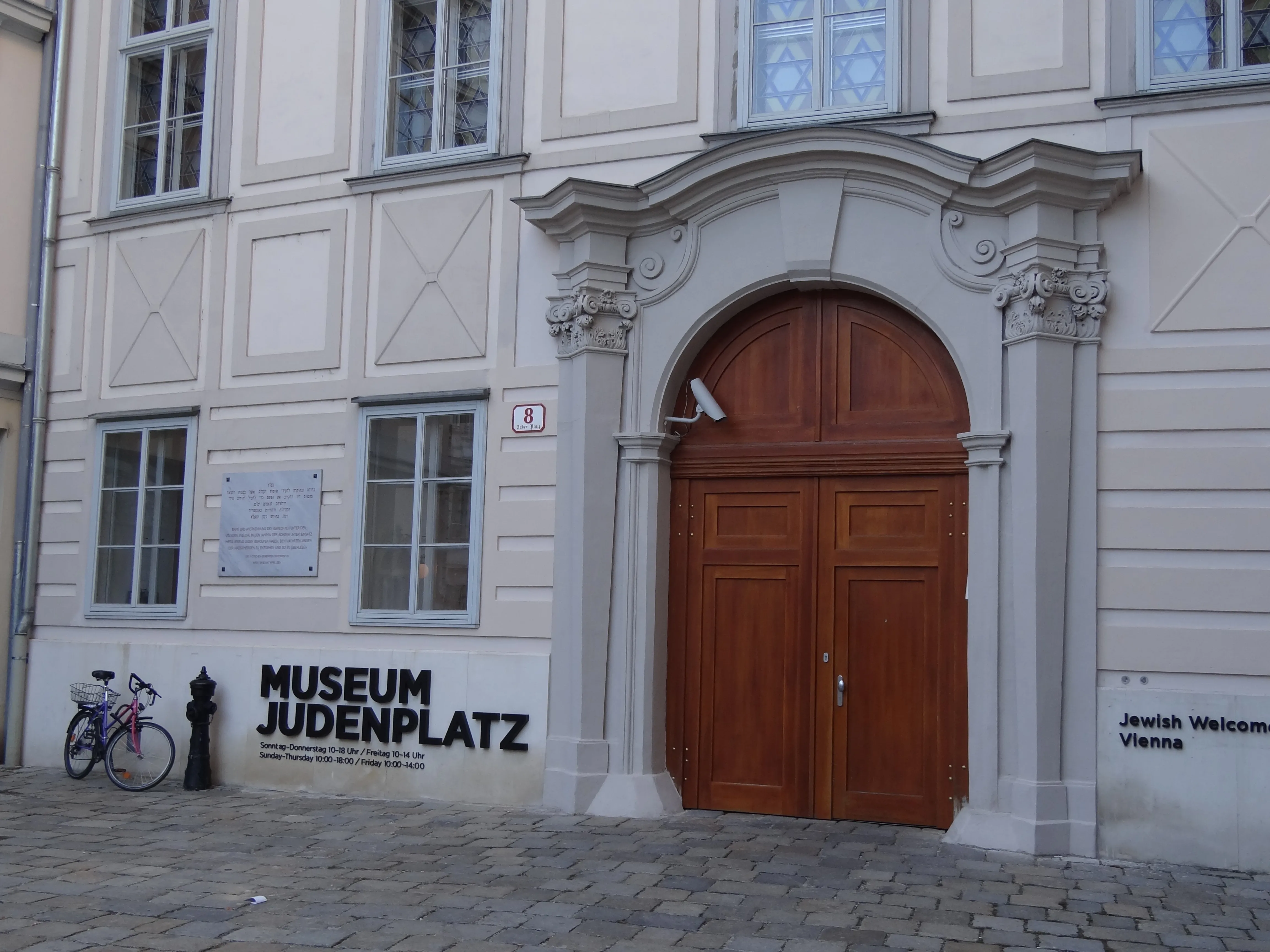 De buitenkant van het Joods Museum Wenen op de Judenplatz, met een grote houten deur, stenen boog en een bord aan de muur van het gebouw.