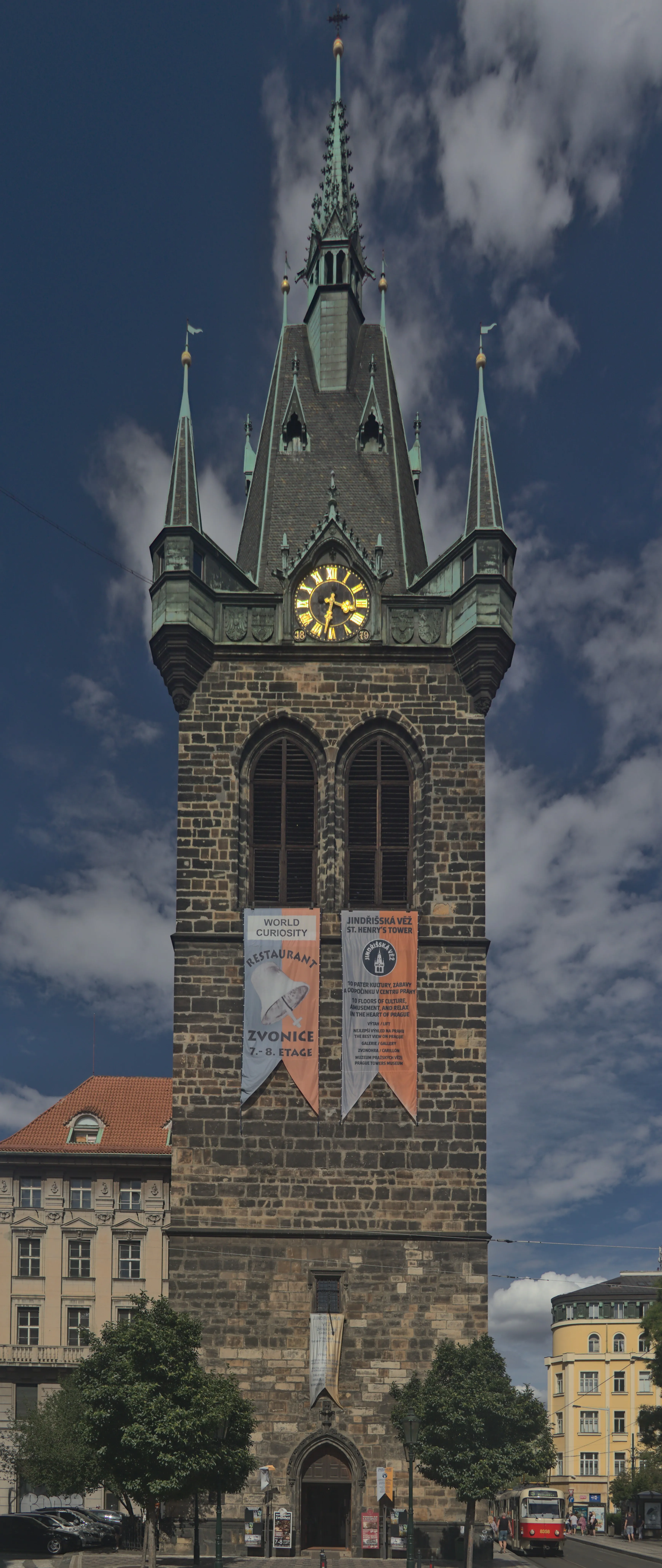 Hoge gotische toren met een klok en groene torenspitsen, stenen gevel en banieren, tegen een blauwe lucht met wolken