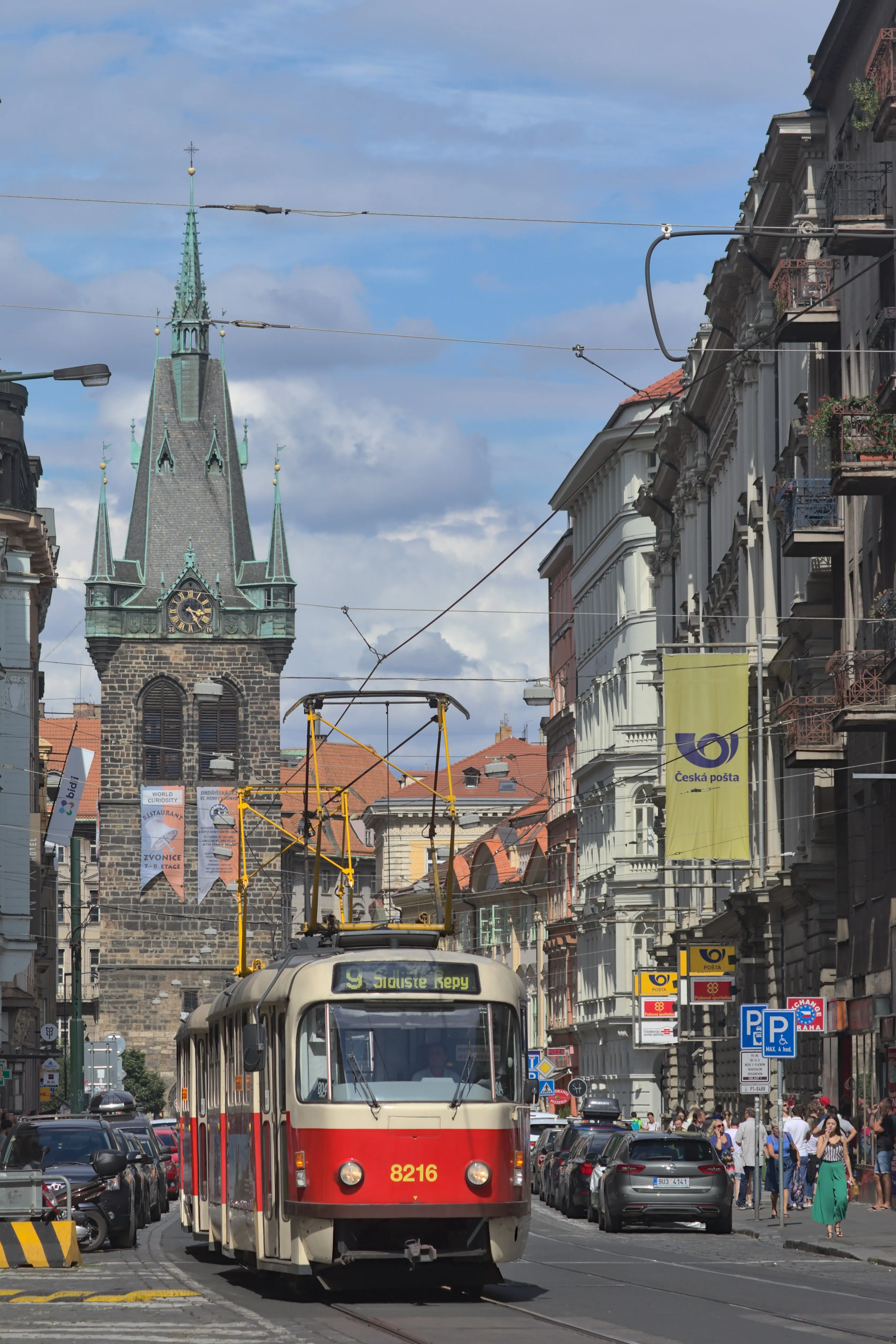 Een rode en crème tram in een stadsstraat met de Jindřišská Toren op de achtergrond.