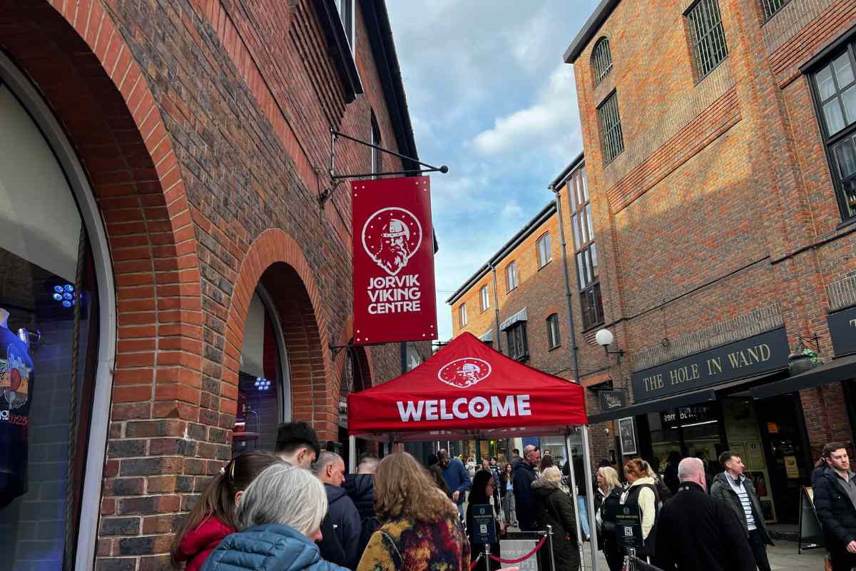 Zewnątrz budynek z czerwonej cegły z wiszącym banerem JORVIK Viking Centre oraz czerwonym zadaszeniem przy wejściu, z odwiedzającymi stojącymi w kolejce na zewnątrz.