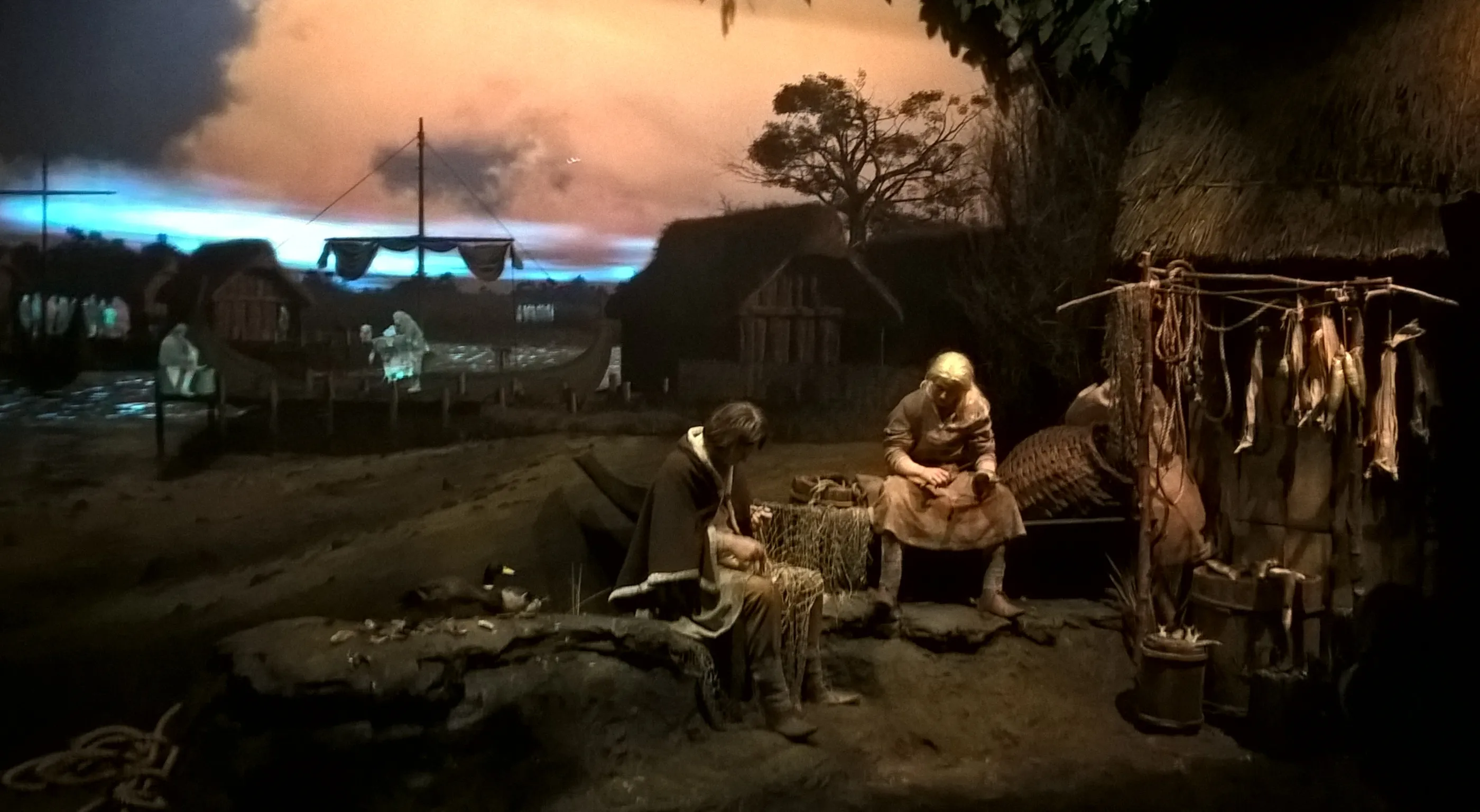 Słabo oświetlona, zrekonstruowana sceneria ulicy z epoki wikingów z manekinami pracującymi przy palenisku i suszoną żywnością, na tle namalowanej panoramy drewnianych budynków.