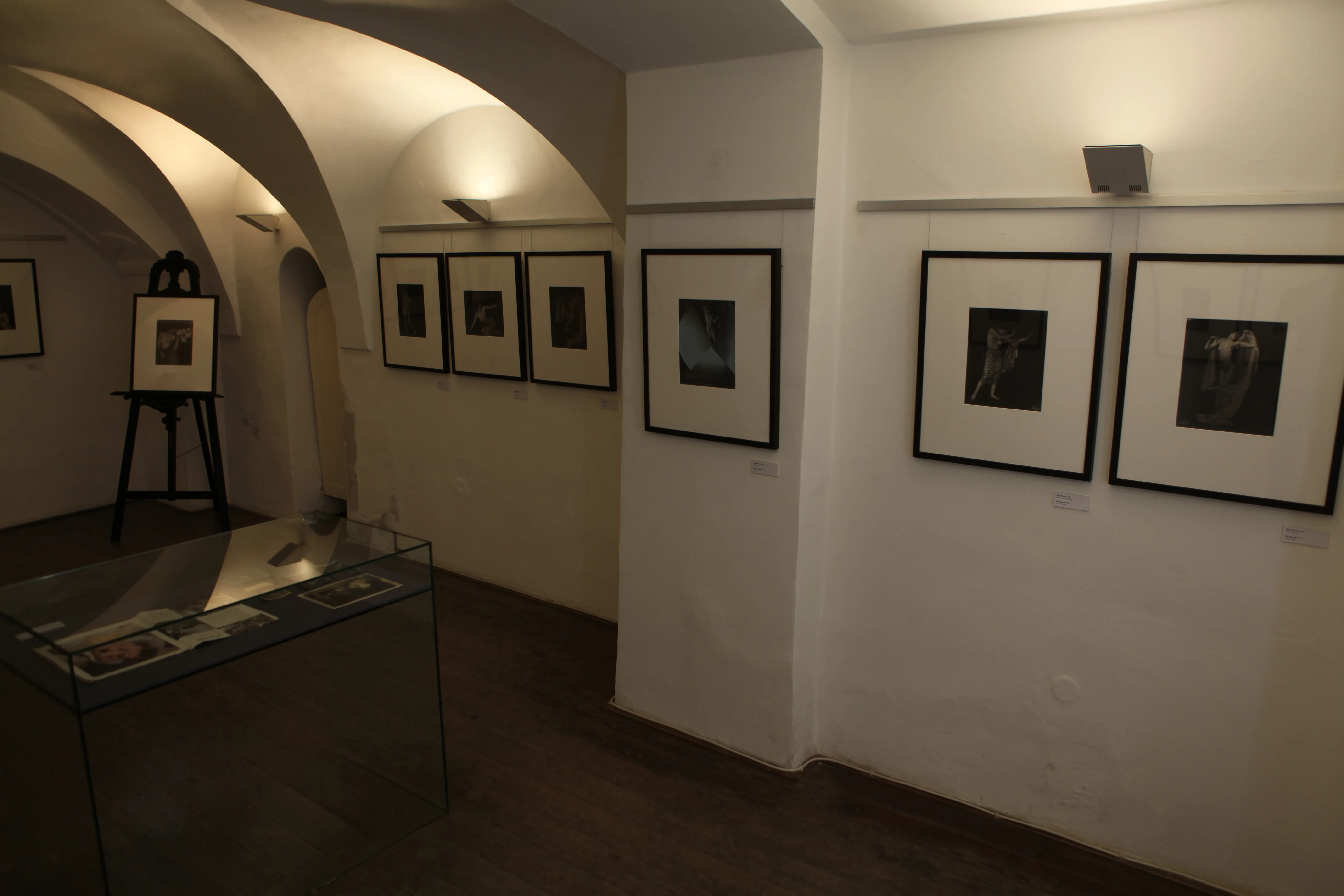 Wnętrze galerii z łukowatymi sufitami, białymi ścianami i oprawionymi fotografiami wystawionymi na sprzedaż.