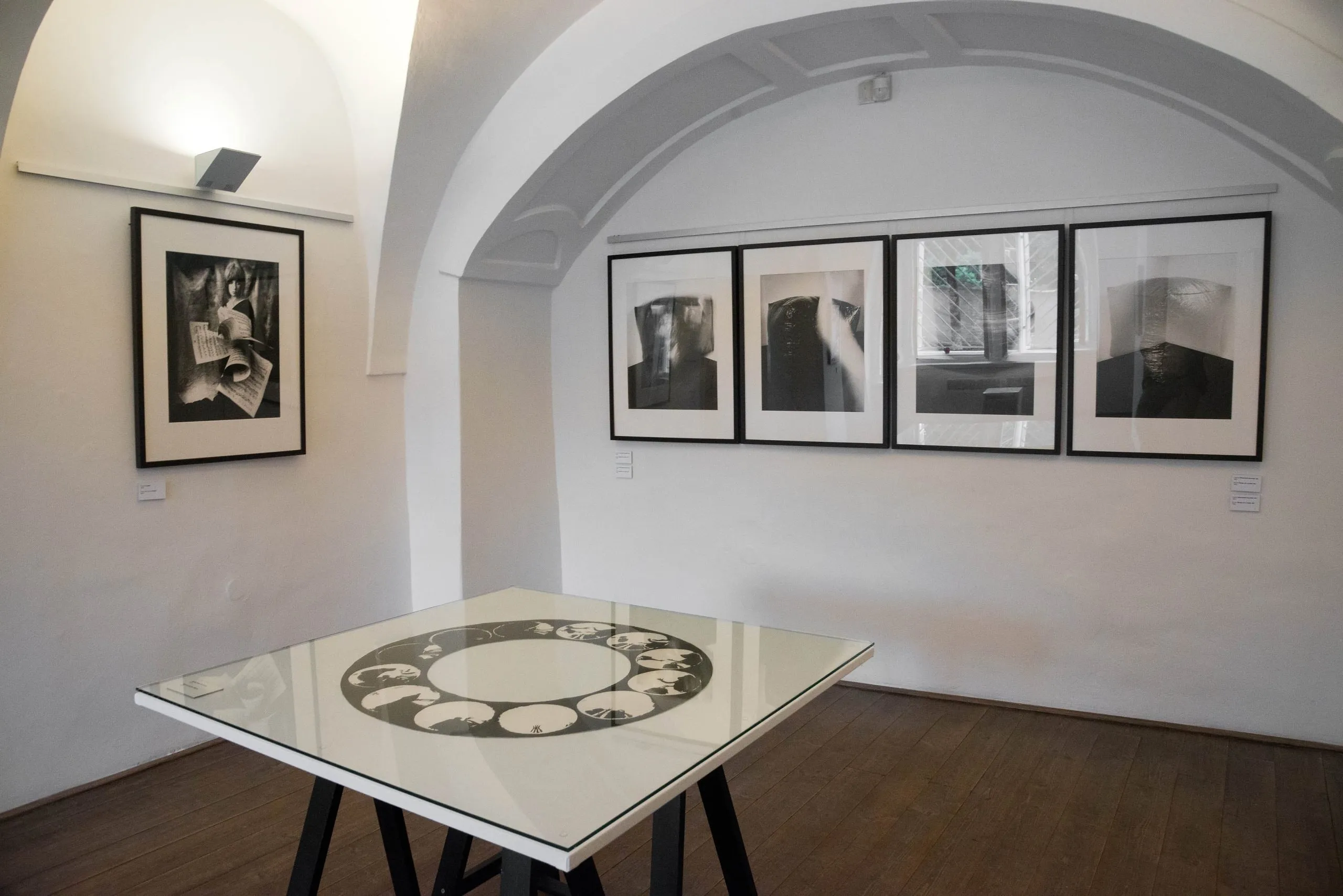 Pomieszczenie galerii z łukowymi sufitami, białymi ścianami i oprawionymi czarno-białymi fotografiami na wystawie, ze szklanym stołem pośrodku.