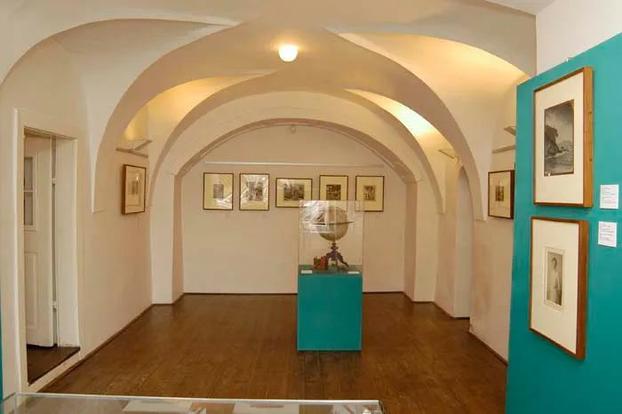 Wnętrze Galerii Josefa Sudka z łukowatymi sufitami, oprawionymi w ramy fotografiami na ścianach i turkusową gablotą pośrodku
