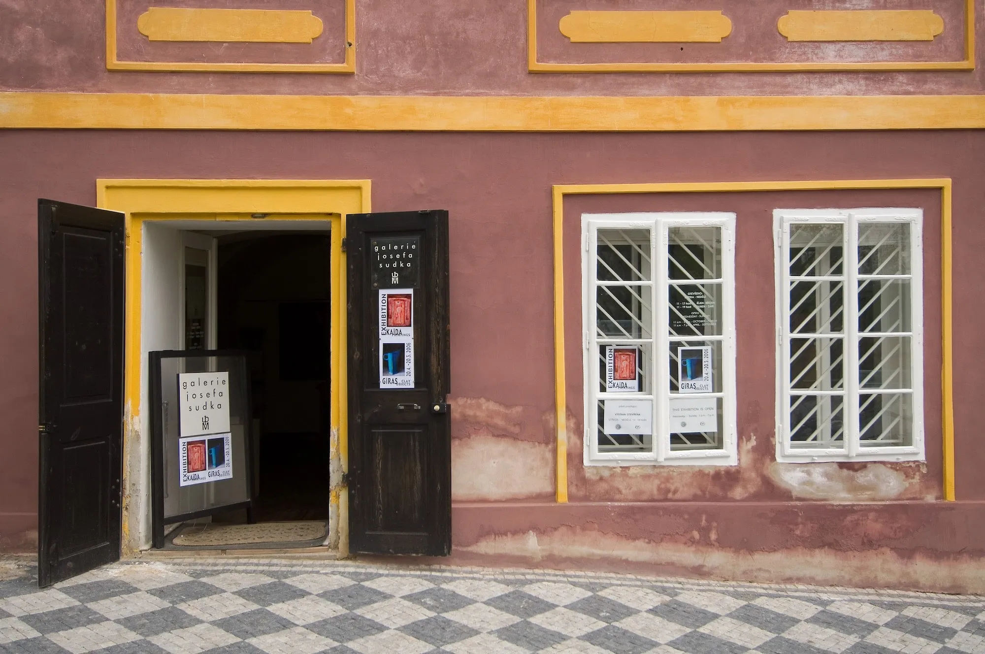 Wejście do budynku z różową fasadą, żółtymi zdobieniami i czarno-białą szachownicową podłogą. Drzwi są otwarte, ukazując znak wewnątrz.