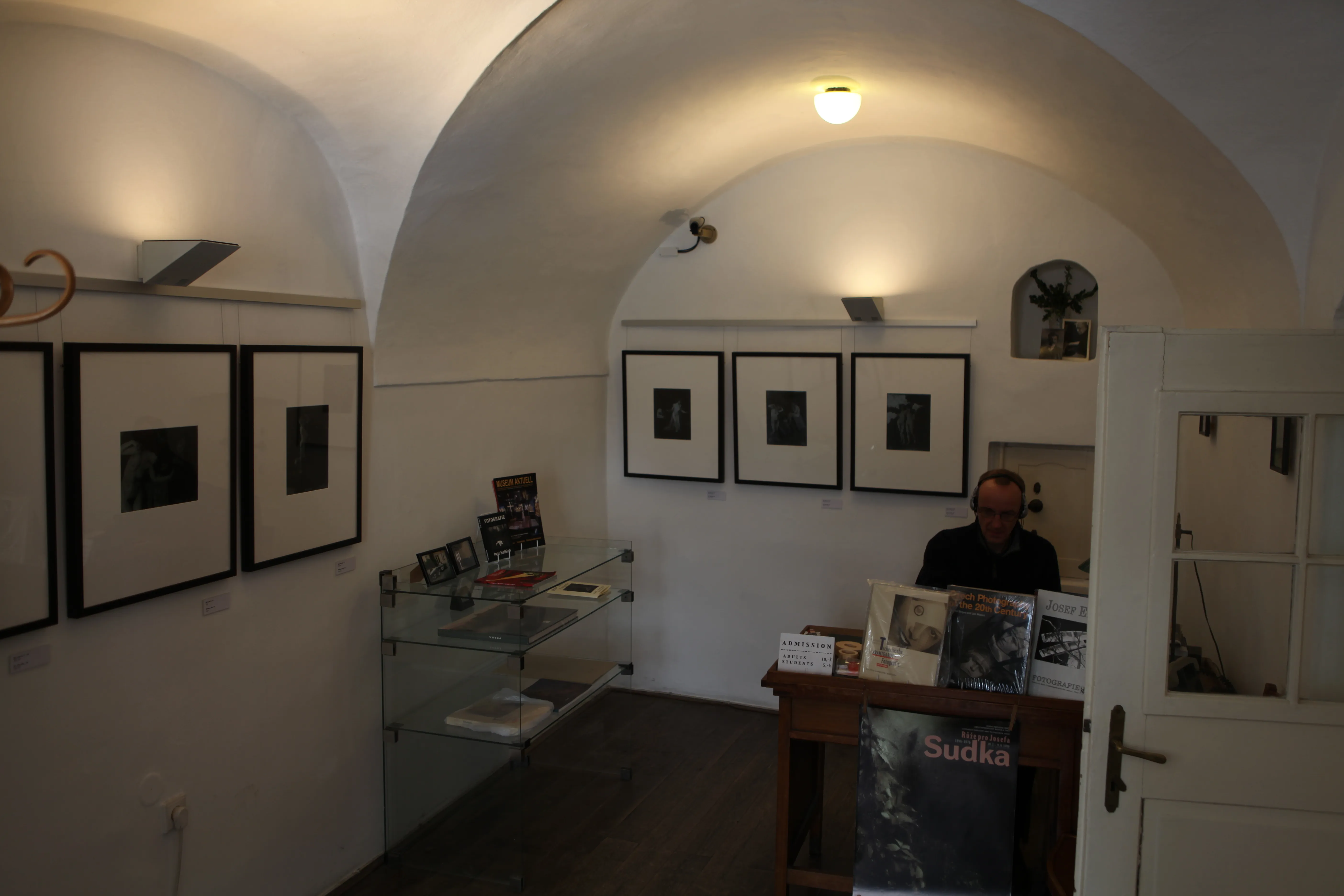 Wnętrze galerii z łukowatymi sufitami, oprawione czarno-białe fotografie na białych ścianach i osoba za ladą recepcyjną.