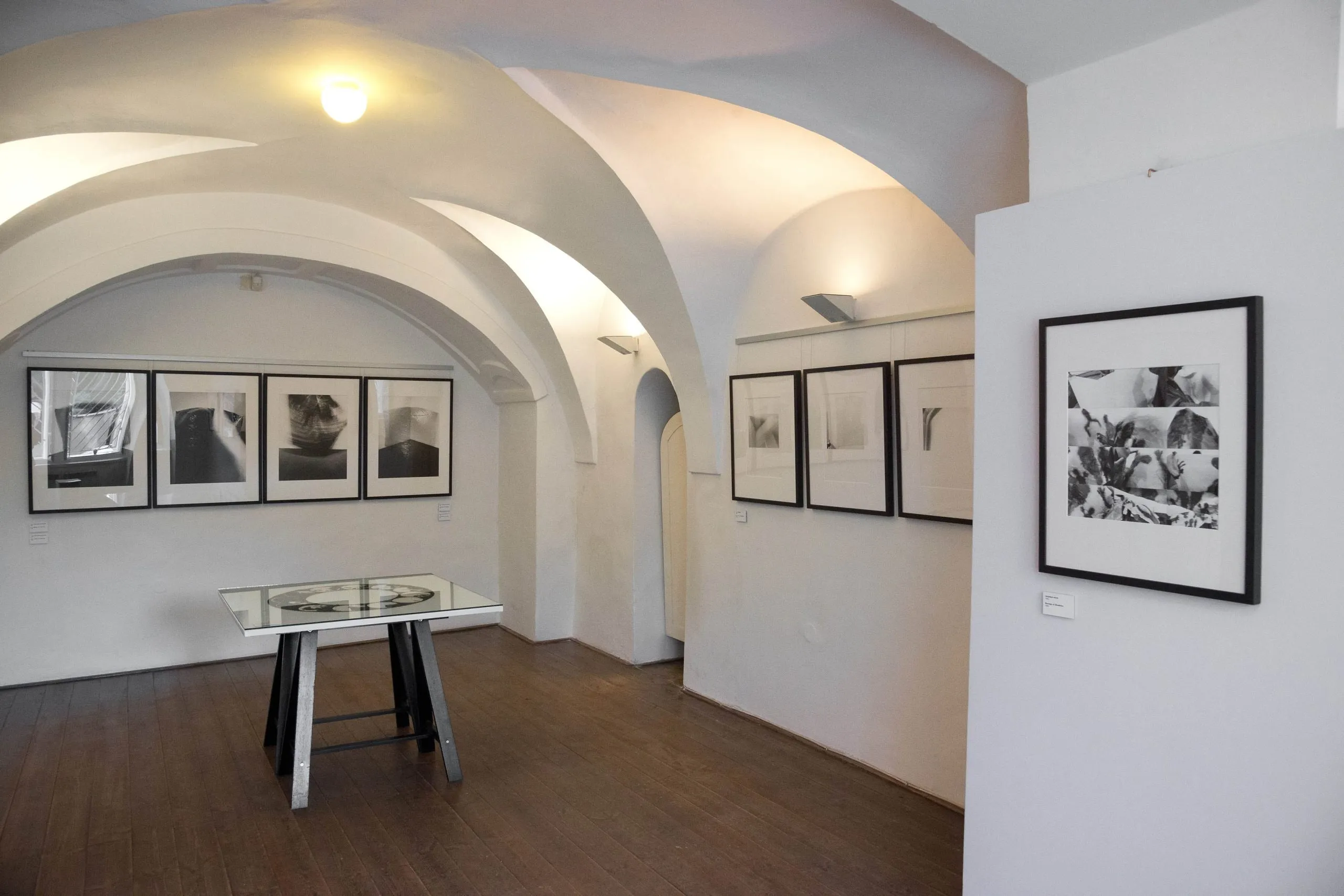 Wnętrze galerii z łukowymi sufitami, białymi ścianami i oprawionymi czarno-białymi fotografiami.