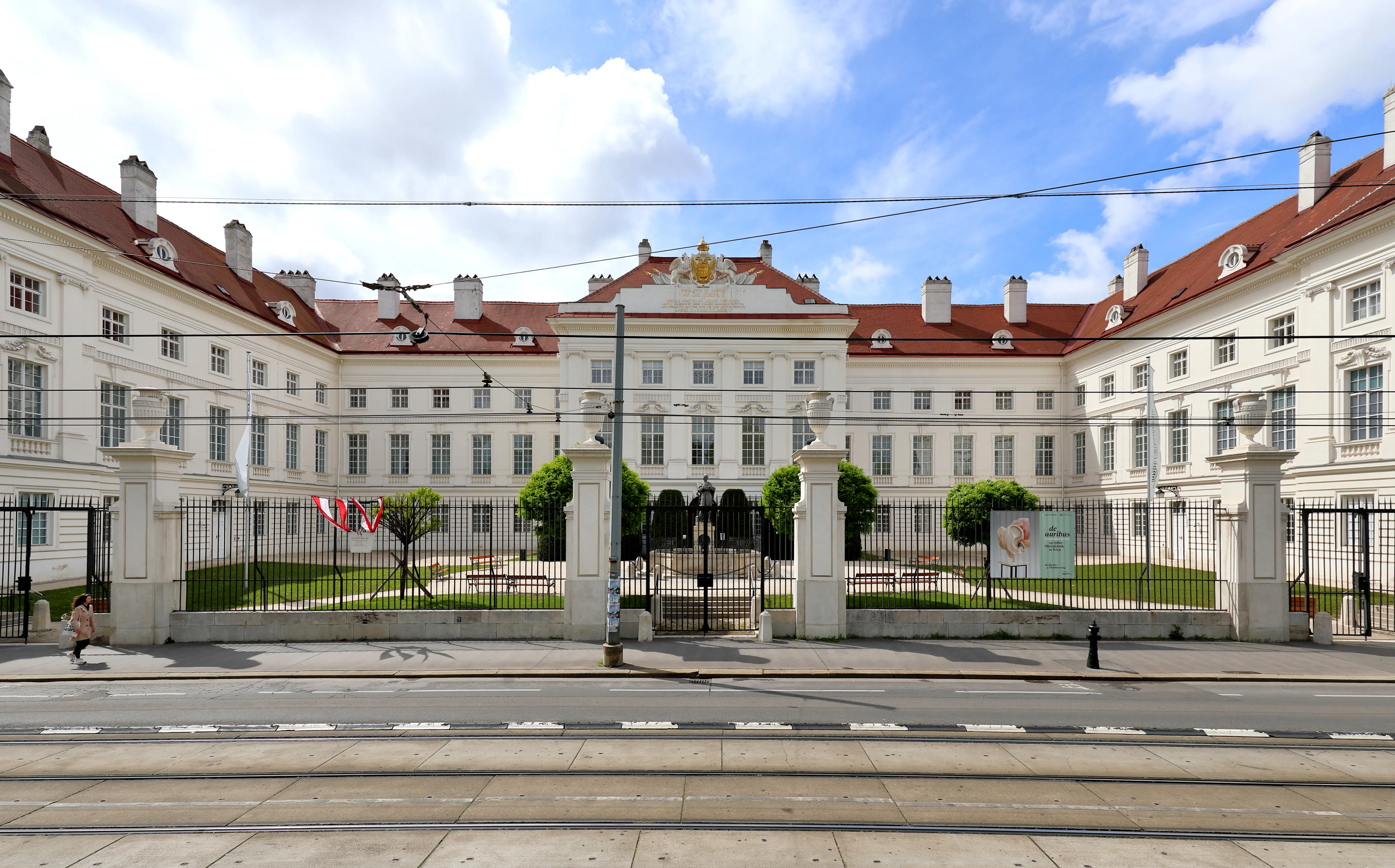 White classical building with red roof and central entrance, surrounded by a black iron fence and green lawn under a blue sky with clouds.