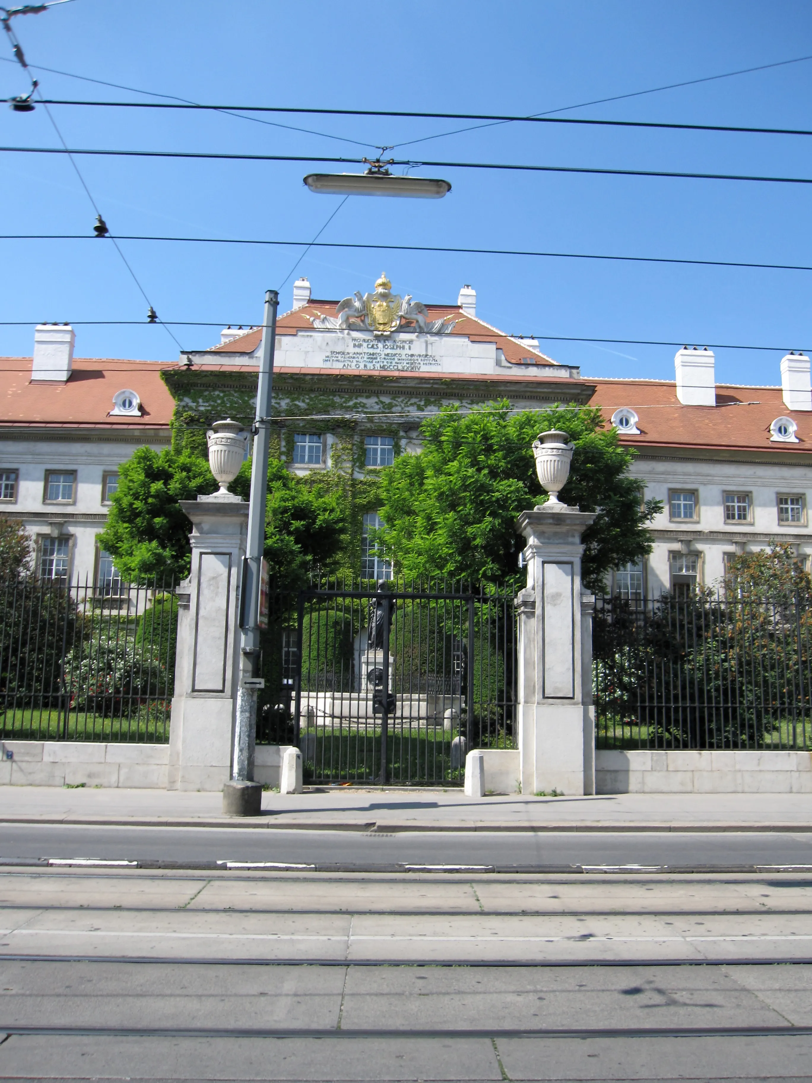 The Josephinum building with a red-tiled roof, white facade, and a central gate with stone pillars and a golden emblem, surrounded by green trees and a black iron fence under a clear blue sky.