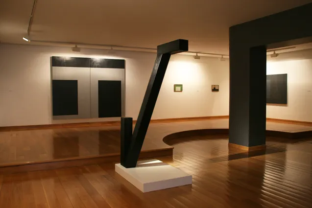 Gallery interior with a large black angular sculpture on a white pedestal, polished wooden floors, white walls with framed artworks, and track lighting