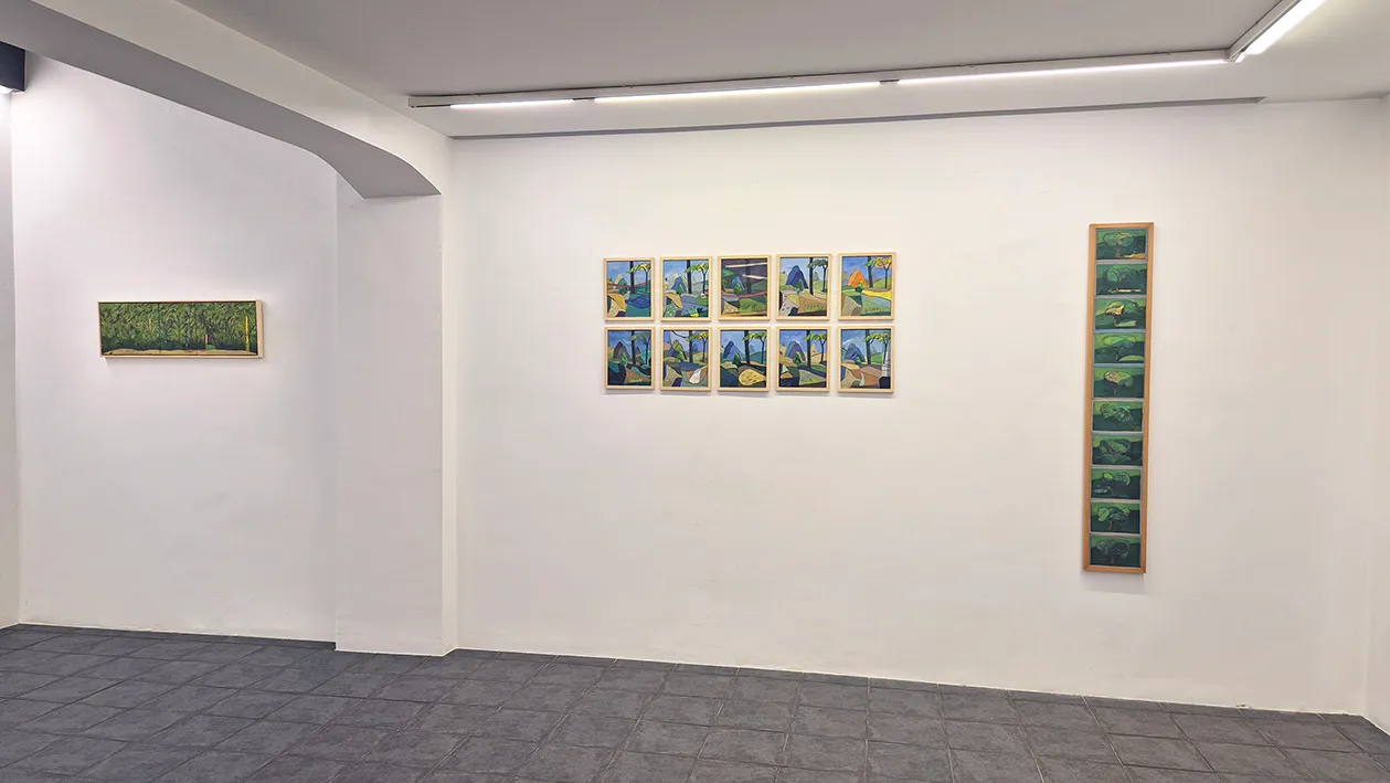 White gallery interior with framed paintings on the walls, including a grid of eight small landscape paintings in the center and a tall vertical painting on the right, under track lighting.