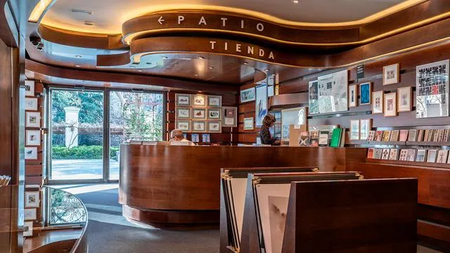 drewniana lada recepcyjna z napisem „PATIO TIENDA” nad nią, oprawione zdjęcia na ścianie i duże okna ukazujące zieleń na zewnątrz