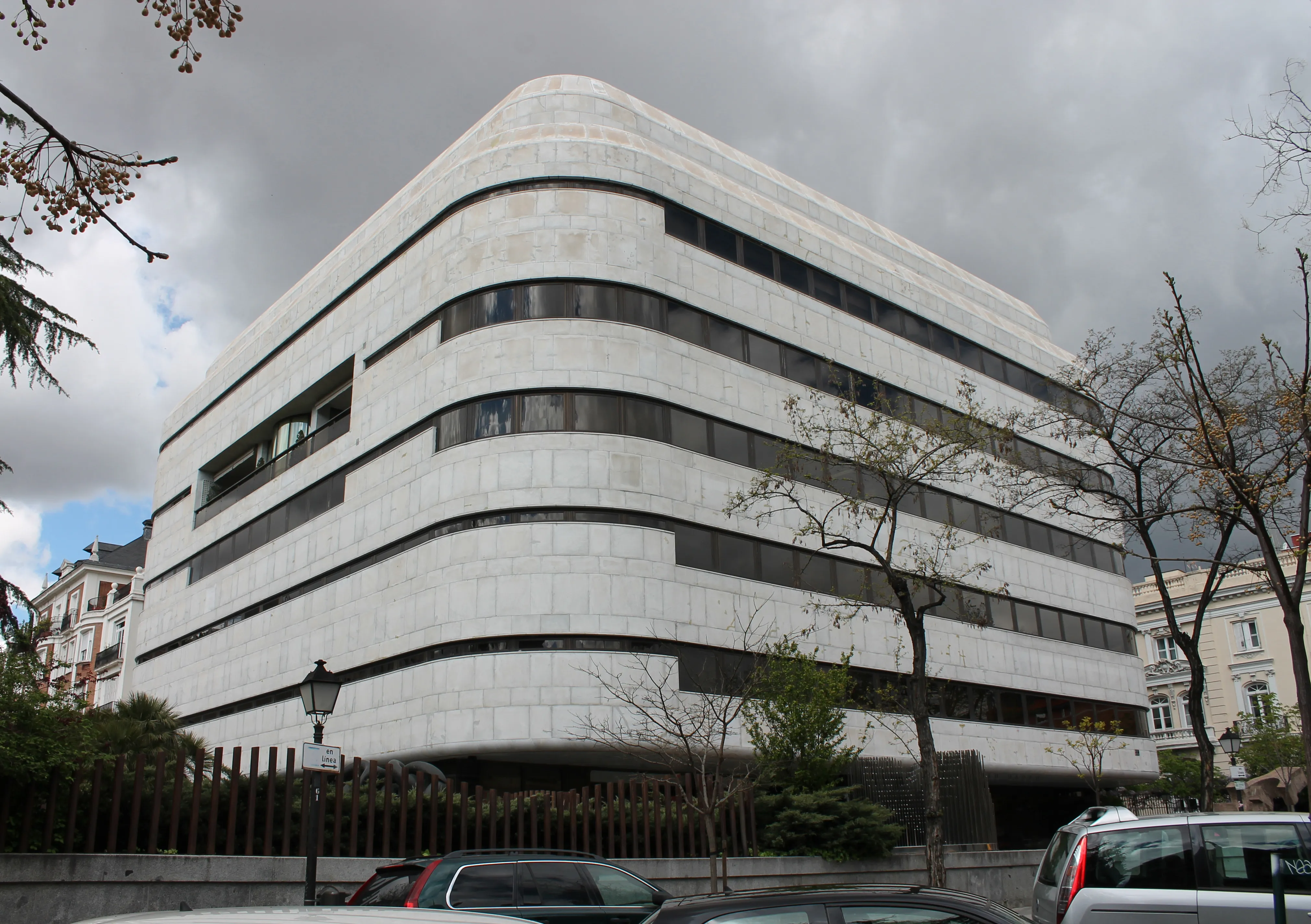 Modern white building with curved facade and horizontal windows, surrounded by trees and parked cars