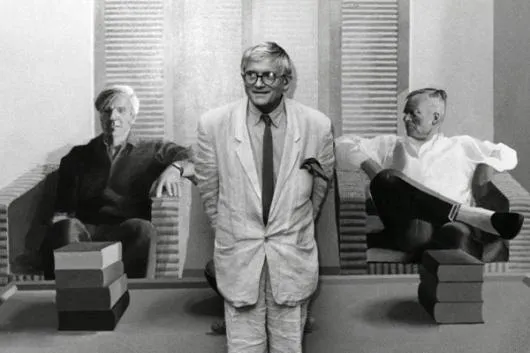 Three men in a gallery, one standing in the center wearing a light suit and glasses, with two others seated on either side, surrounded by stacked boxes and vertical blinds in the background