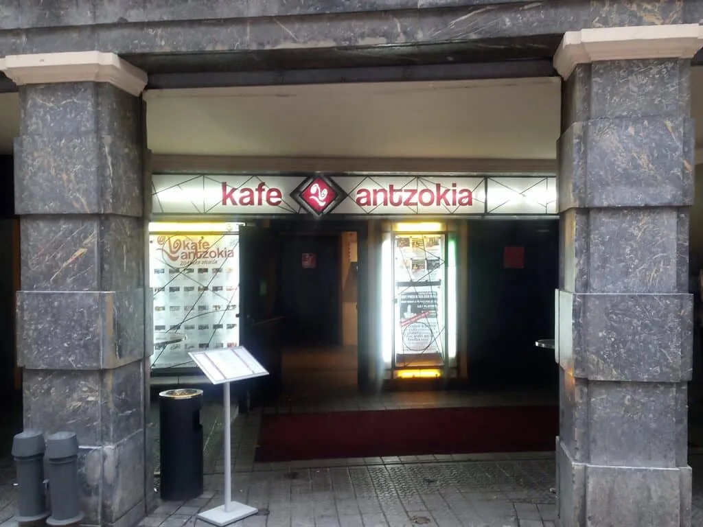 Zewnętrzne wejście do Kafe Antzokia z kamiennymi kolumnami i podświetlanym szyldem nad drzwiami