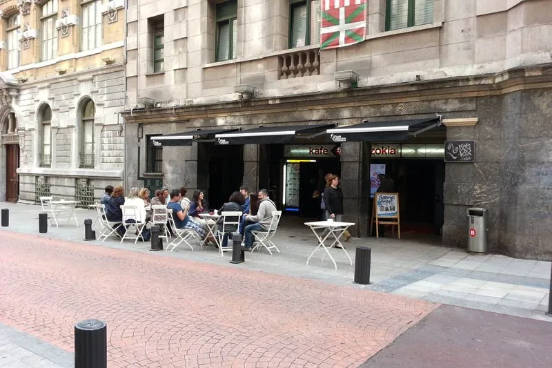 Widok z zewnątrz na Kafe Antzokia z ludźmi siedzącymi przy stolikach na zewnątrz i flagą baskijską wiszącą nad wejściem.
