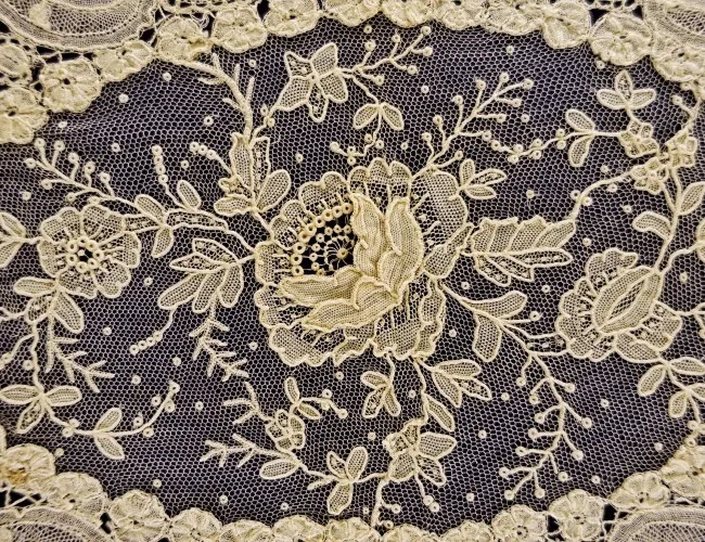 Primer plano de un intrincado encaje floral de color crema con motivos de rosas y enredaderas sobre un fondo oscuro, que muestra detalles de costura y bordes festoneados.