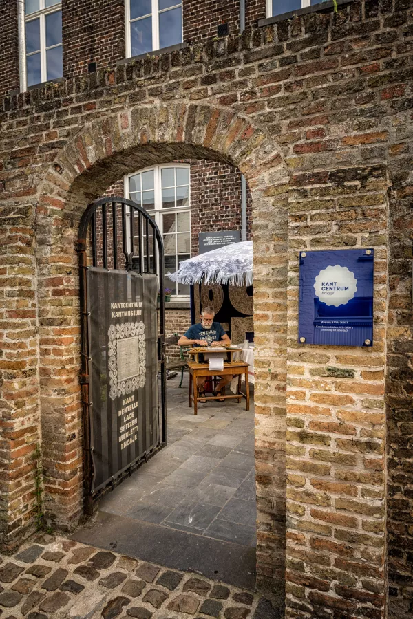 Entrada de arco de ladrillo con puerta de metal y letrero azul de Kantcentrum, con una encajera trabajando en una mesa visible a través de la puerta.