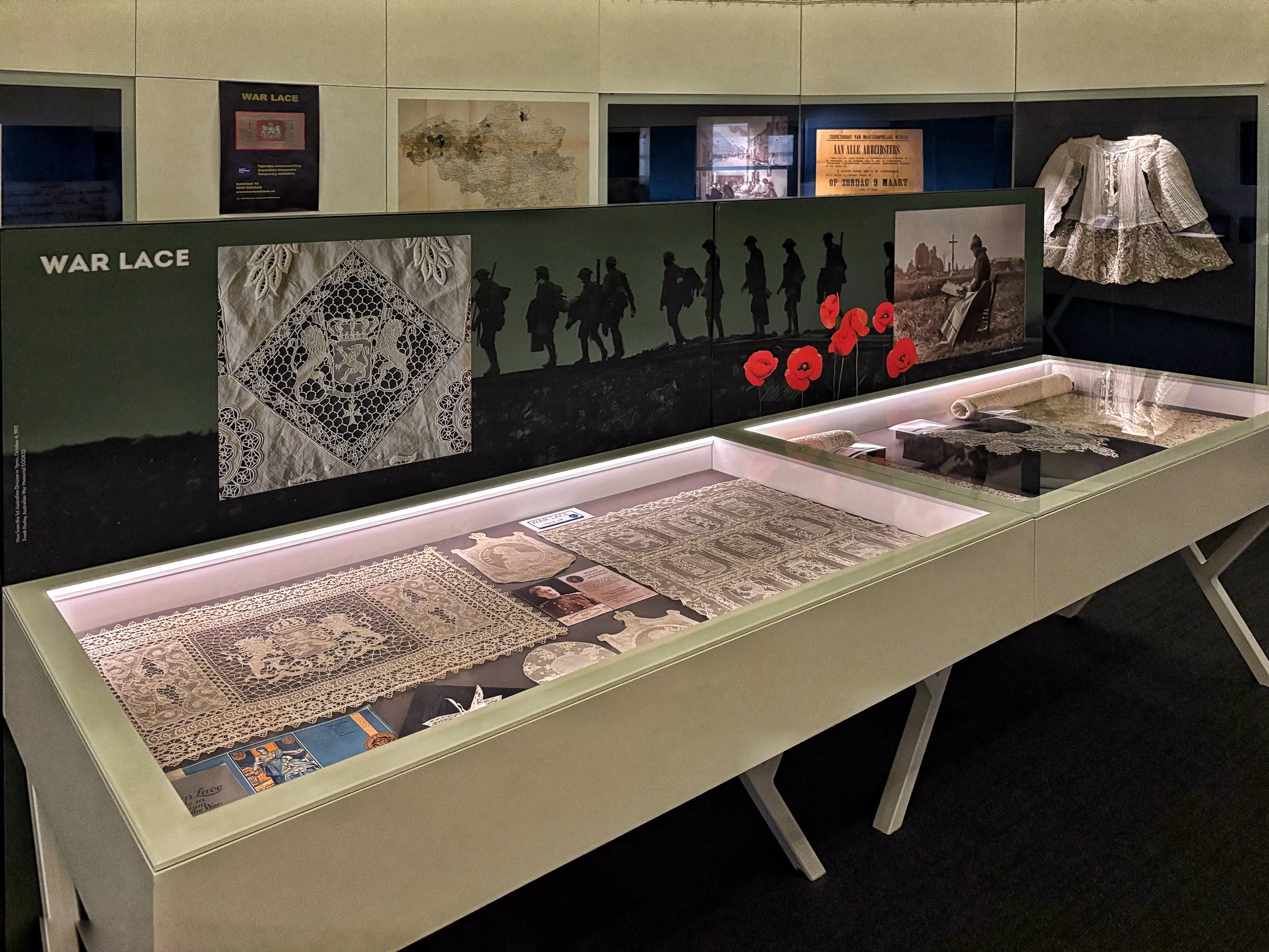 Vitrina de cristal con piezas de encaje histórico y, detrás, un panel de la exposición 'War Lace' que muestra siluetas de soldados y amapolas rojas