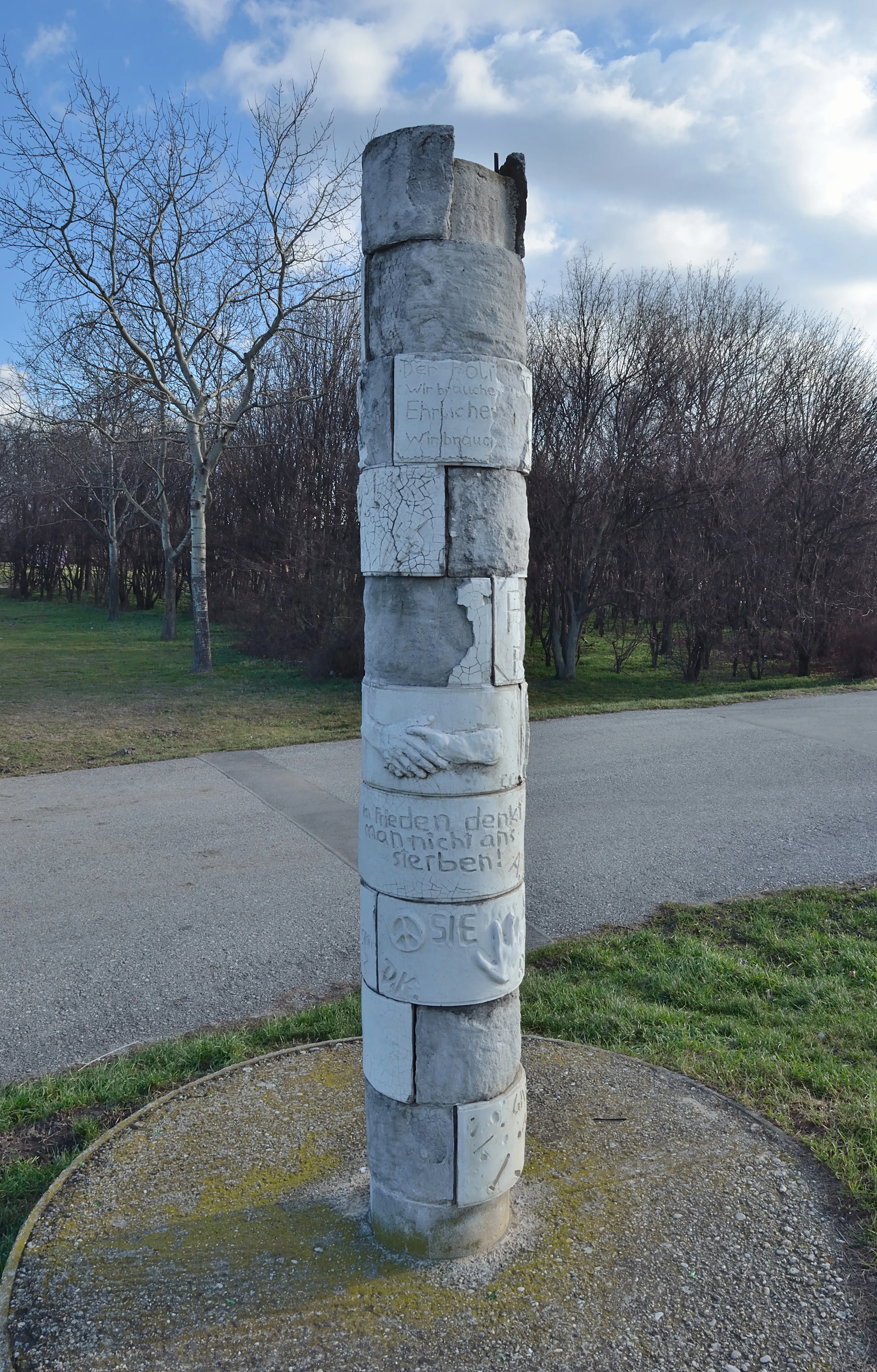 A stone memorial pillar with engraved text and carvings, standing in a grassy area with trees and a path behind it.