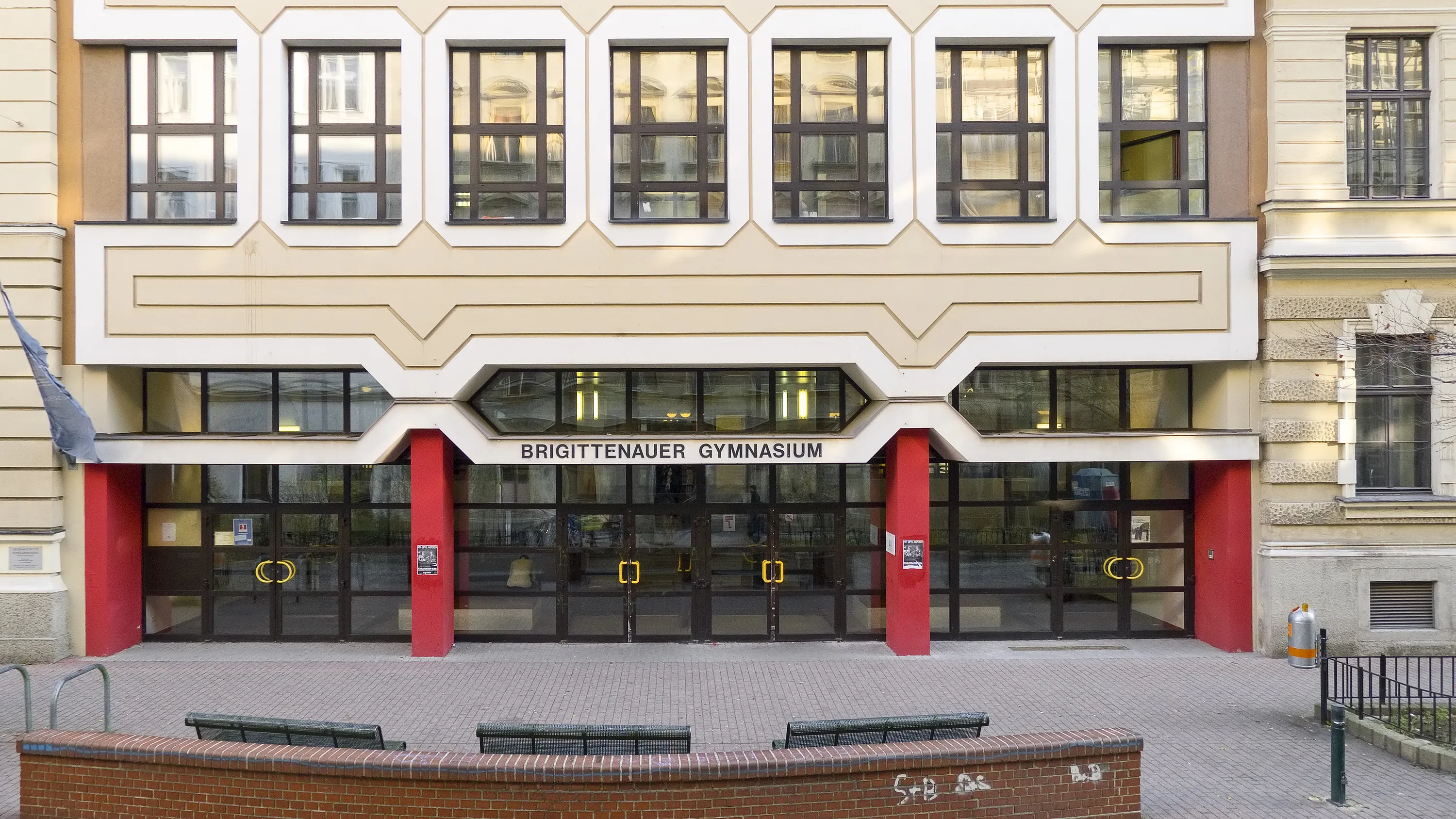 The facade of a school building with a sign reading 'BRIGITTENAUER GYMNASIUM' above the entrance, featuring large windows and red pillars.