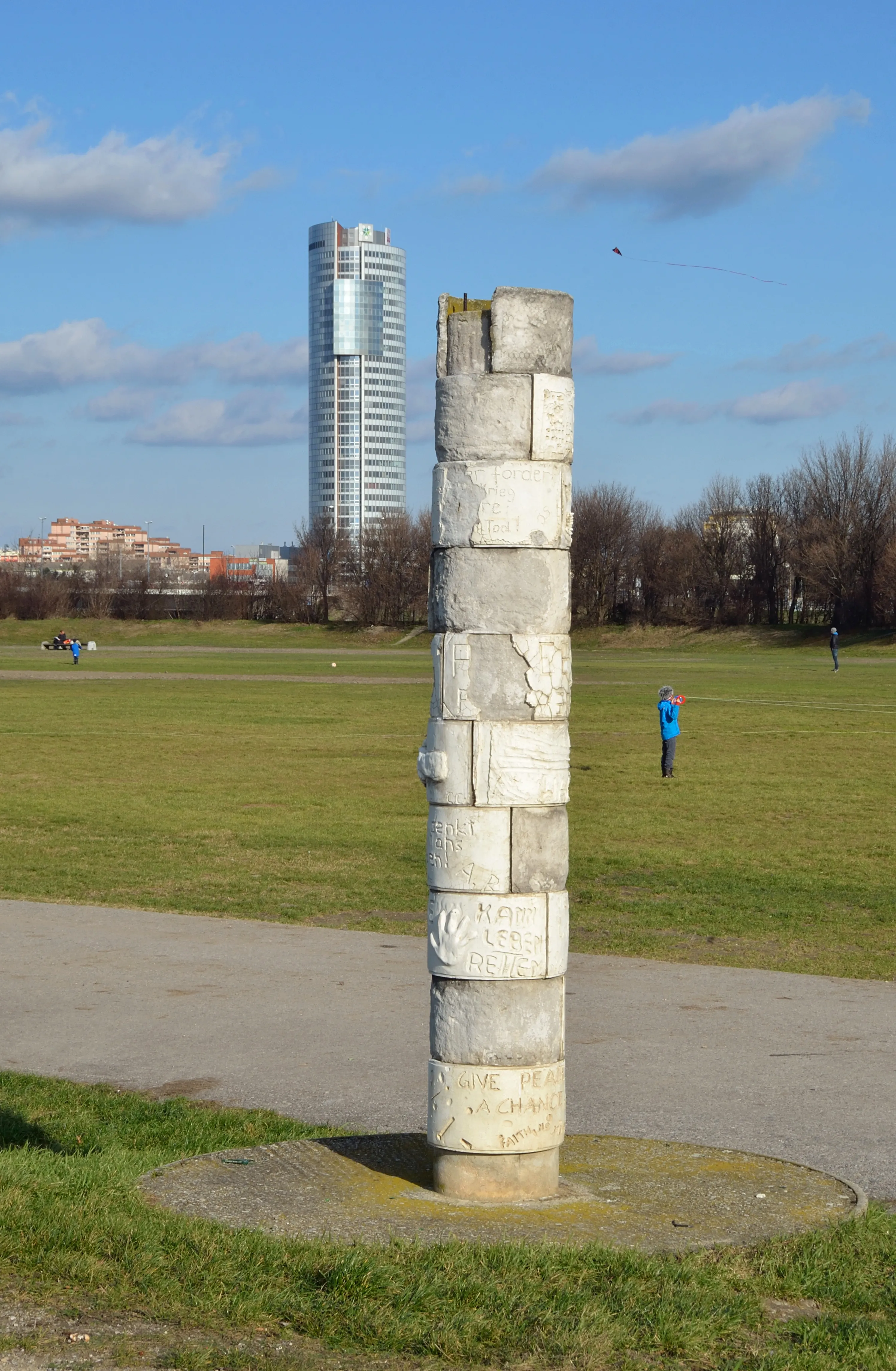 A tall stone monument composed of stacked blocks with inscriptions, situated in a grassy field with a modern building in the background.