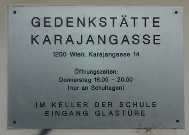 Una placa metálica con texto negro que dice GEDENKSTÄTTE KARAJANGASSE, 1200 Wien, Karajangasse 14, Horario: Jueves 16.00 - 20.00 (solo en días escolares), EN EL SÓTANO DE LA ESCUELA ENTRADA PUERTA DE CRISTAL