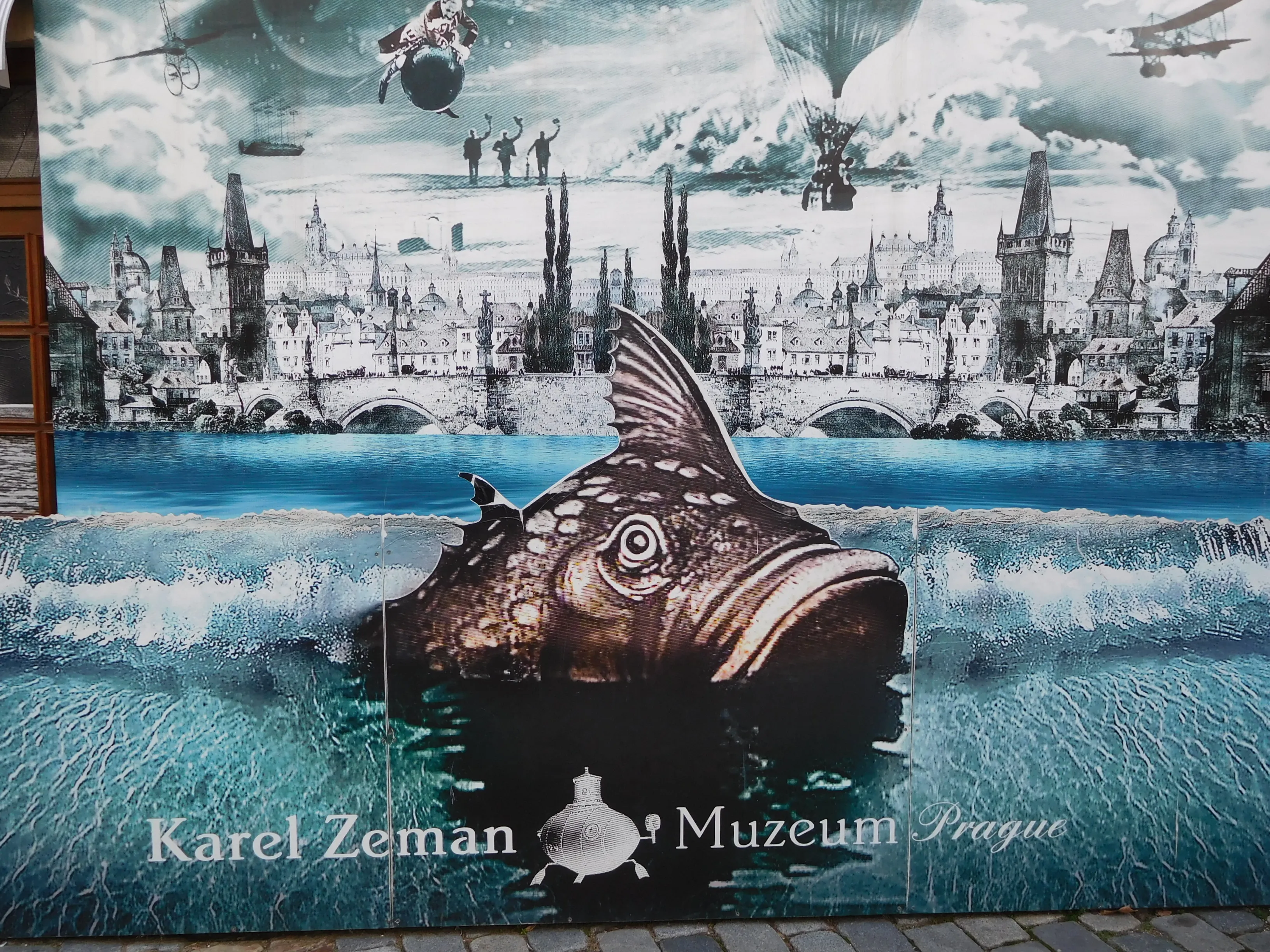 A large mural on a building wall showing a fantastical scene with a giant fish, a cityscape with a bridge, and flying machines, with the text 'Karel Zeman Museum Prague' at the bottom.