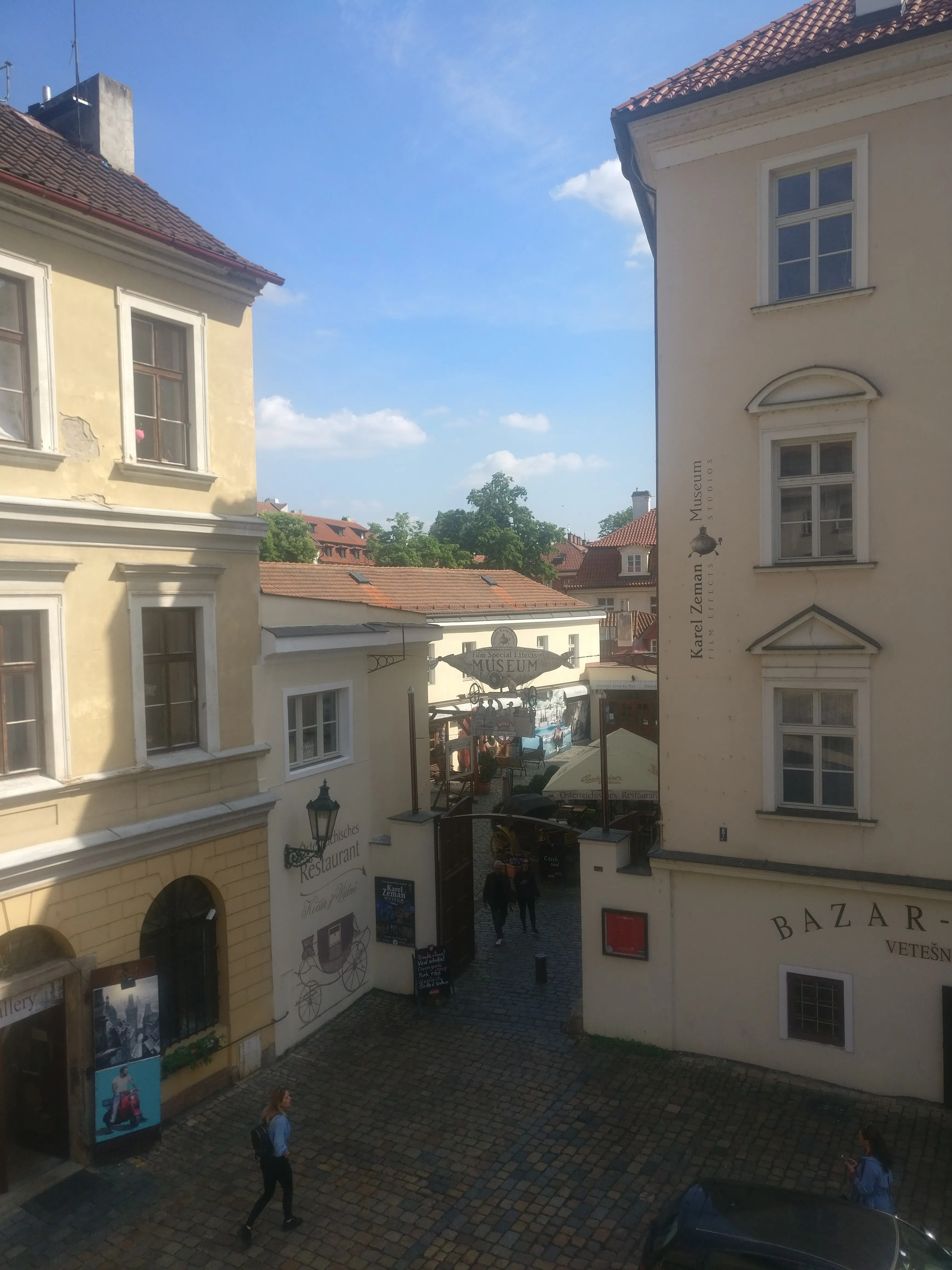 Cobblestone street with historic buildings, a sign for the Karel Zeman Museum, and people walking on a sunny day.