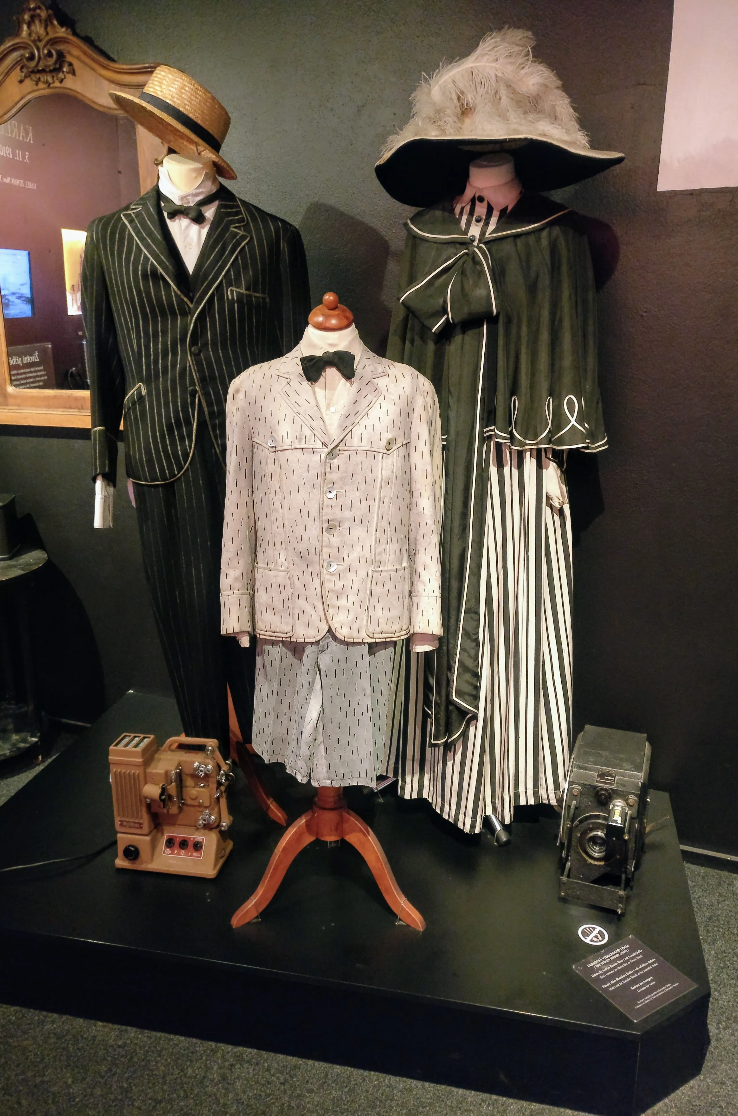 Three mannequins dressed in vintage costumes, including a pinstripe suit, a patterned suit, and a long green coat with a large hat, displayed with old film cameras on a black platform.