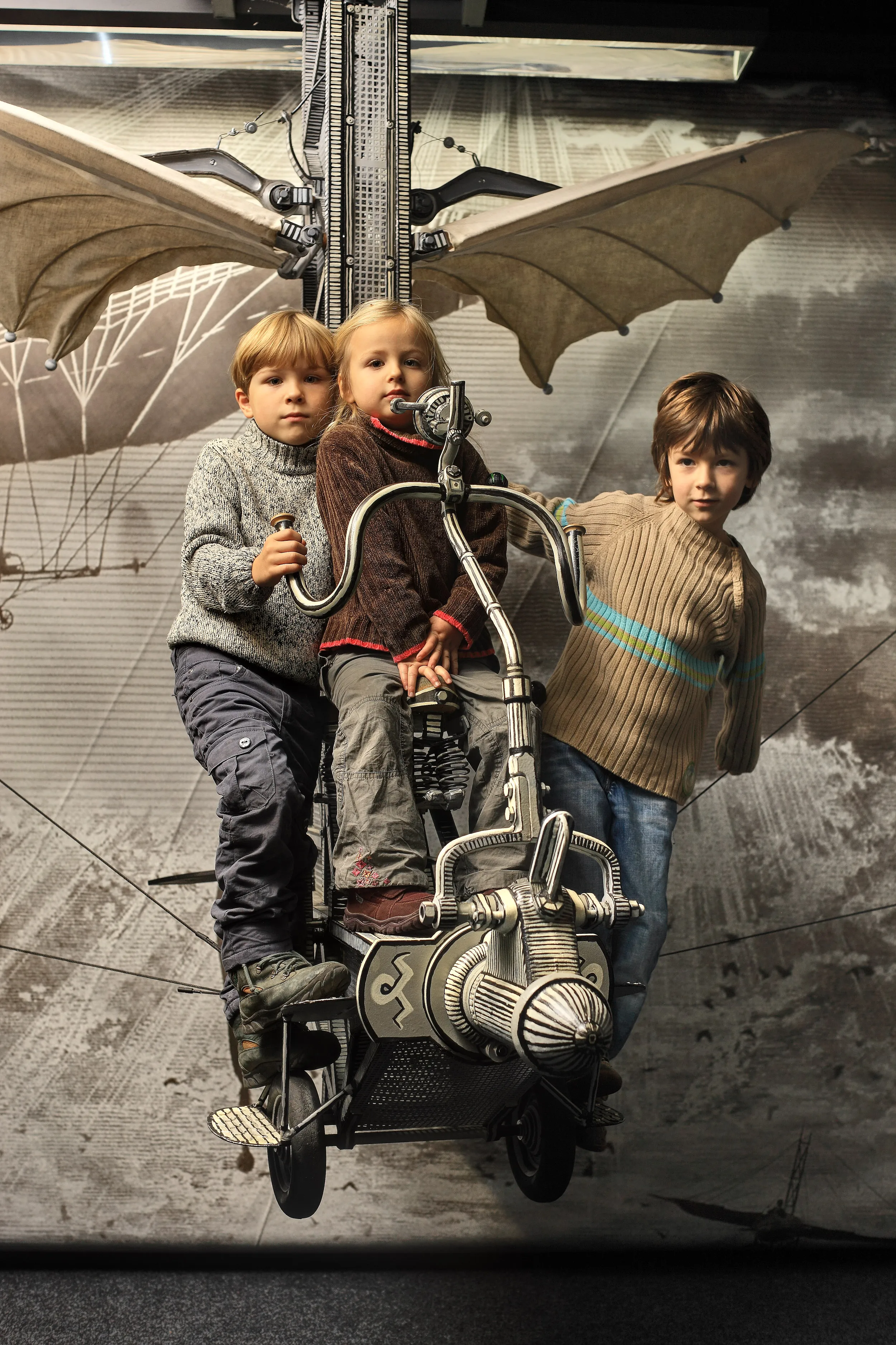 Three children sitting on a mechanical flying machine with bat wings in an exhibition space