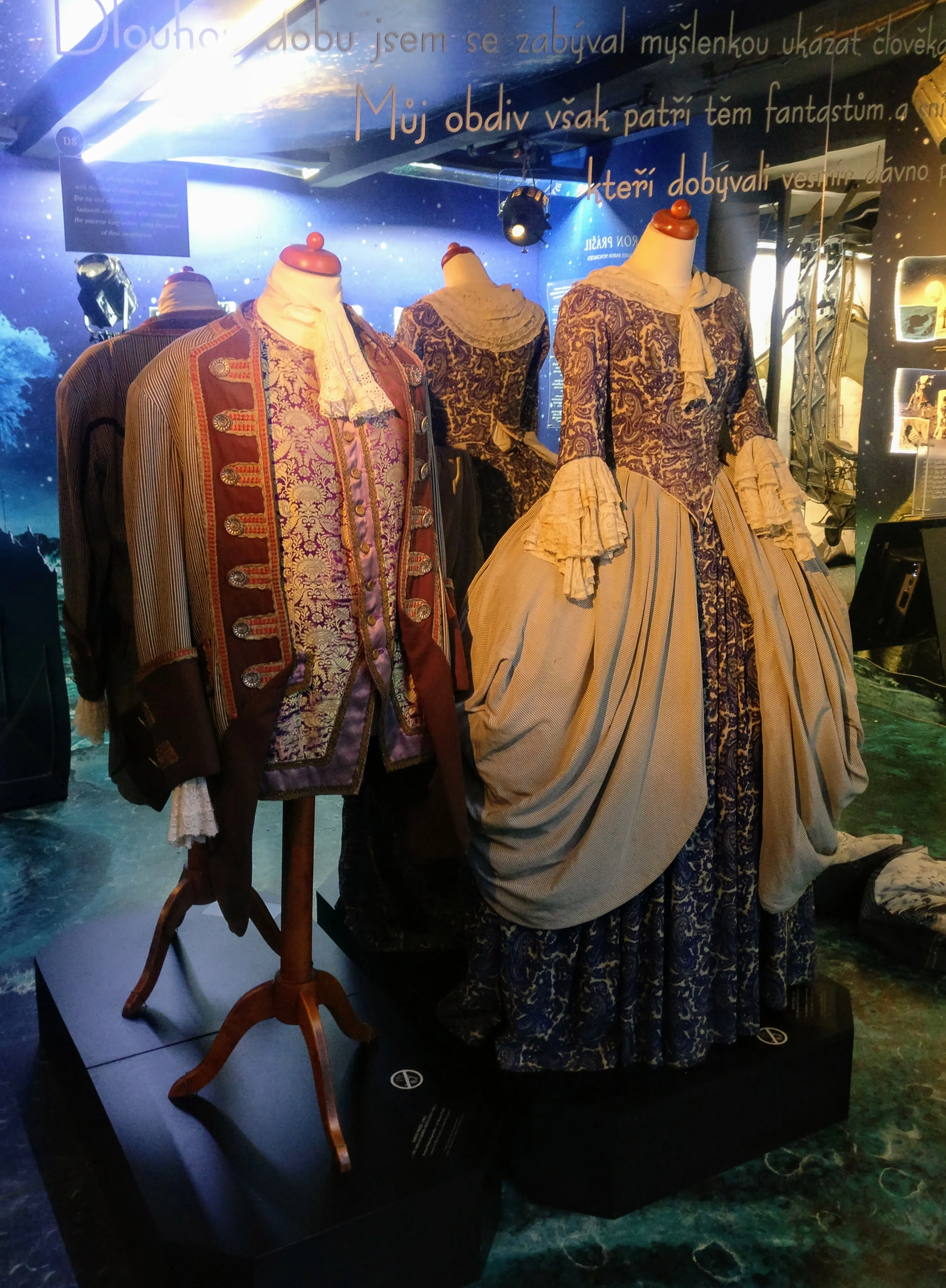 Historical costumes on mannequins in a museum exhibit with Czech text on the wall