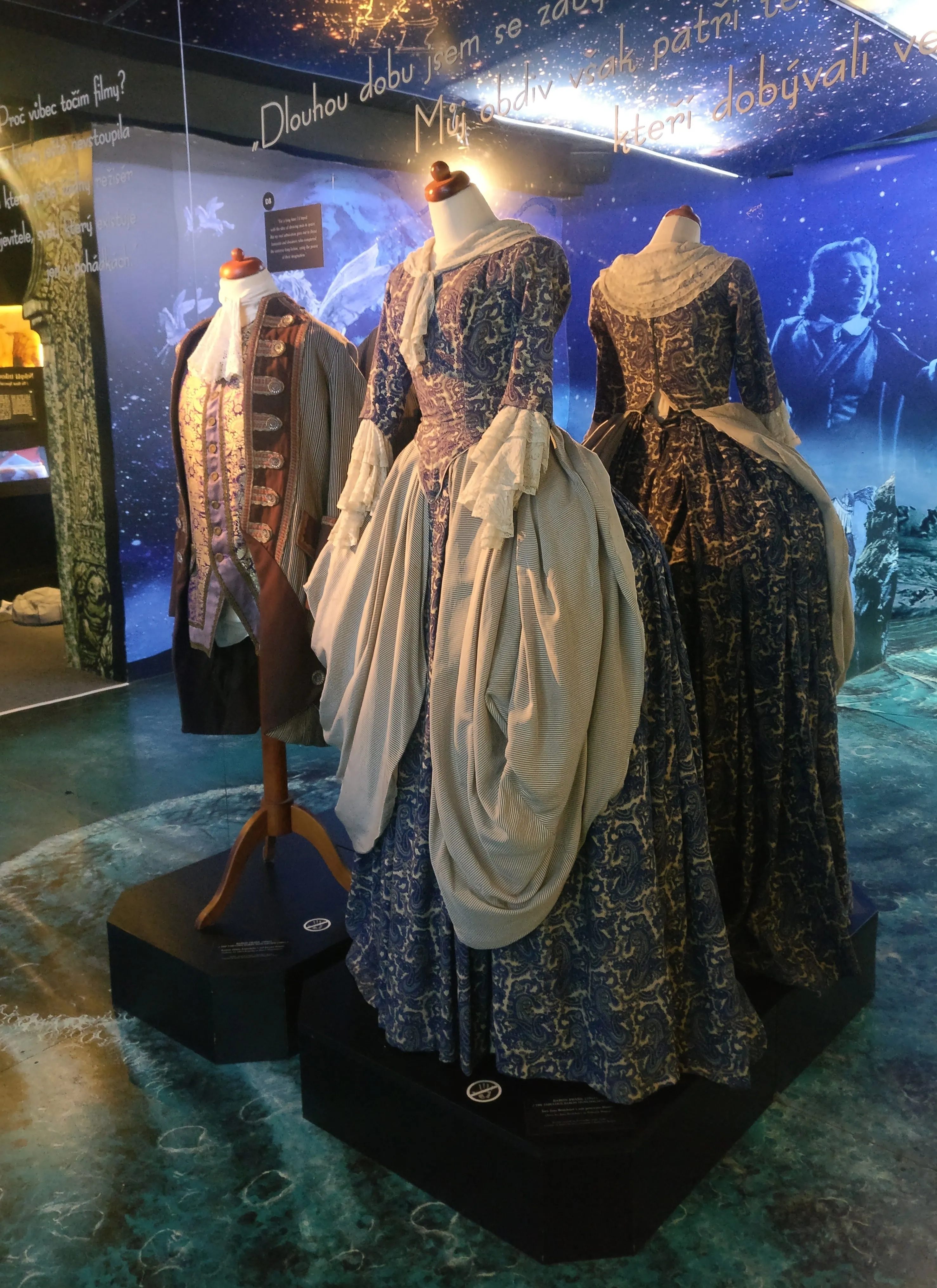 Three mannequins displaying historical costumes with elaborate patterns and layered fabrics in a museum exhibit